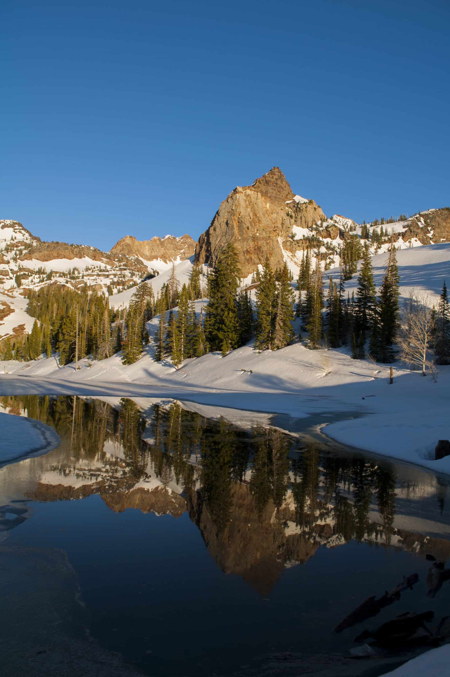 A calm view of Lake Blanche in spring, surrounded by lingering snow, evergreens and a partial reflection under a crisp blue sky.