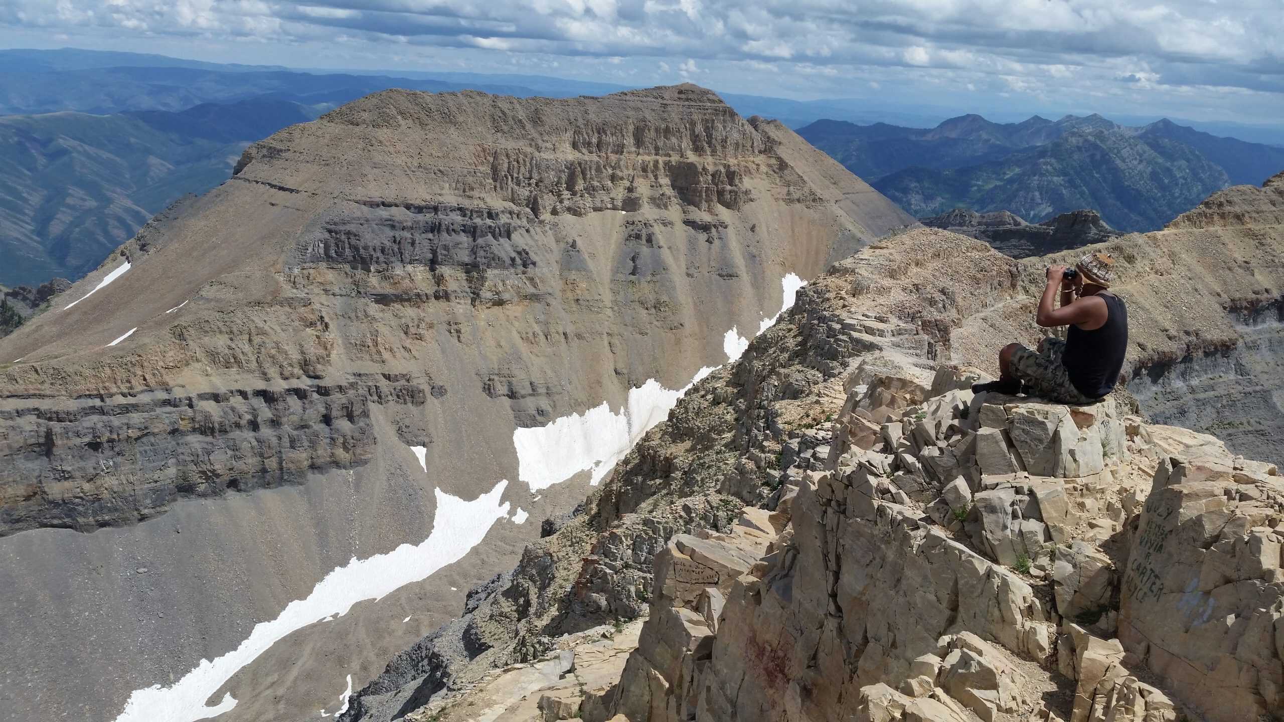 A hiker sits on the rocky edge of Mount Timpanogos, looking out over a dramatic landscape of layered cliffs and distant peaks, truly a surreal hike in Utah. 