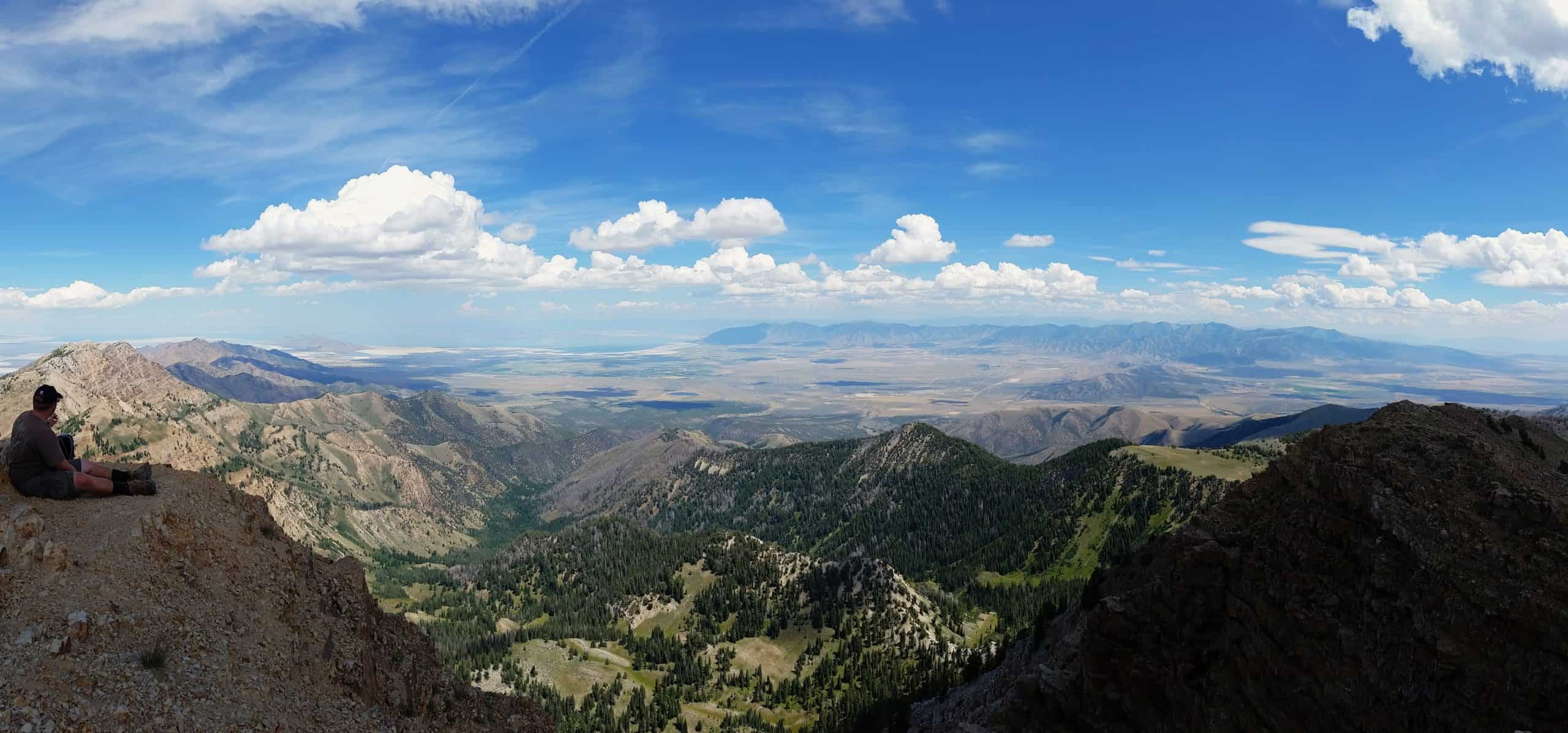 A hiker resting on a rocky ridge near the summit of Deseret Peak overlooking a vast desert landscape.