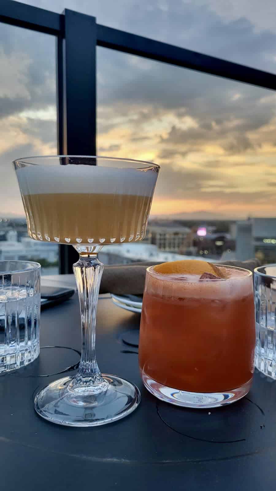 Two cocktails on a rooftop table at sunset, with Salt Lake City and a colorful sky in the background.