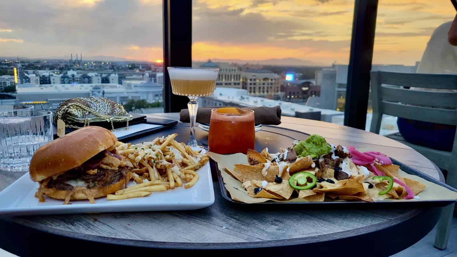 Rooftop dinner spread with a burger, fries, nachos, and cocktails on a table overlooking downtown Salt Lake City at sunset.