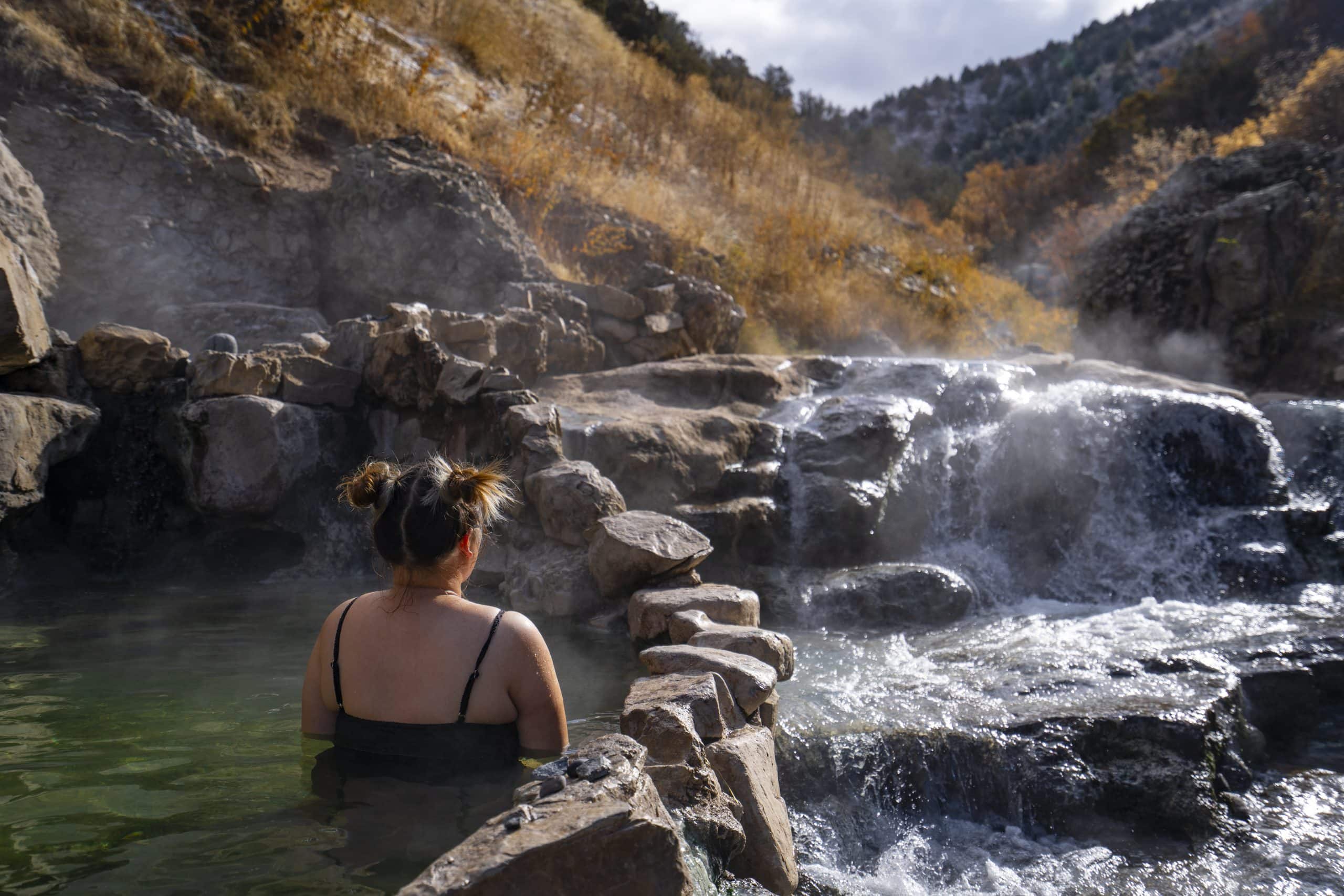 A woman relaxes in a steaming natural pool surrounded by rocky formations and autumn foliage while water cascades over stone ledges nearby. 