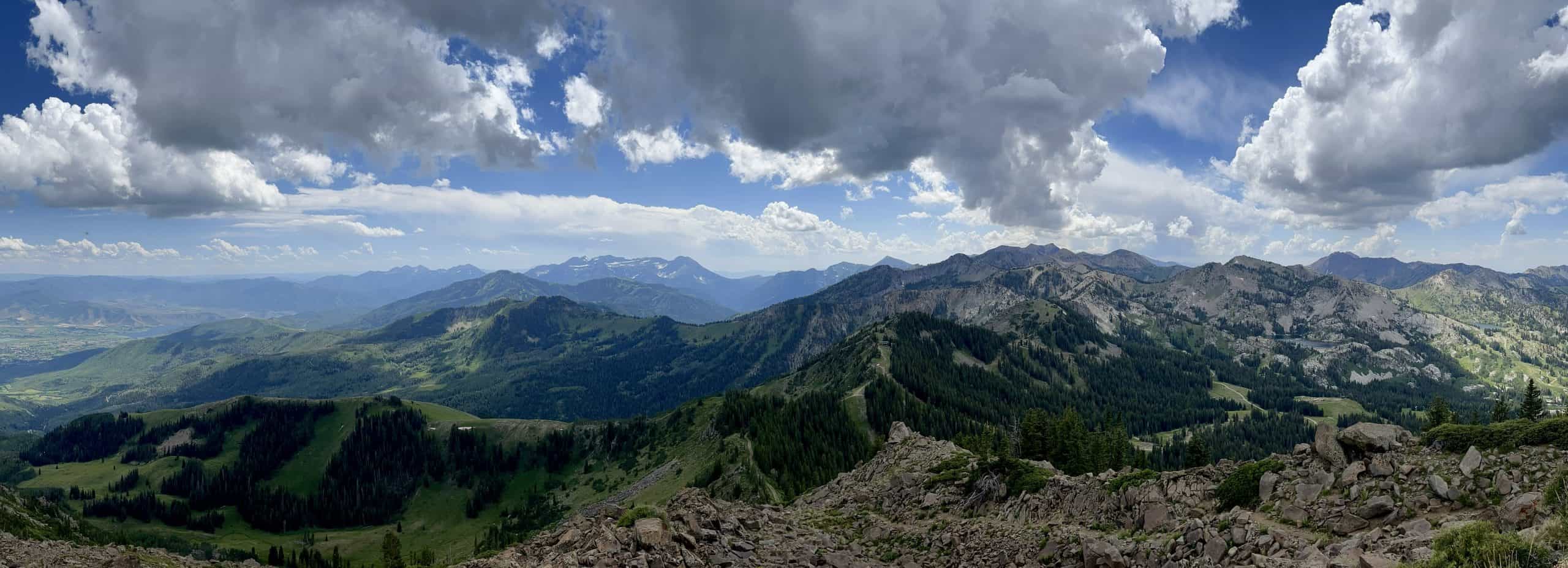 Vast mountain view fromt the Clayton Peak Summit in Big Cottonwood Canyon, with green ridges and distant peaks under cloudy skies. 