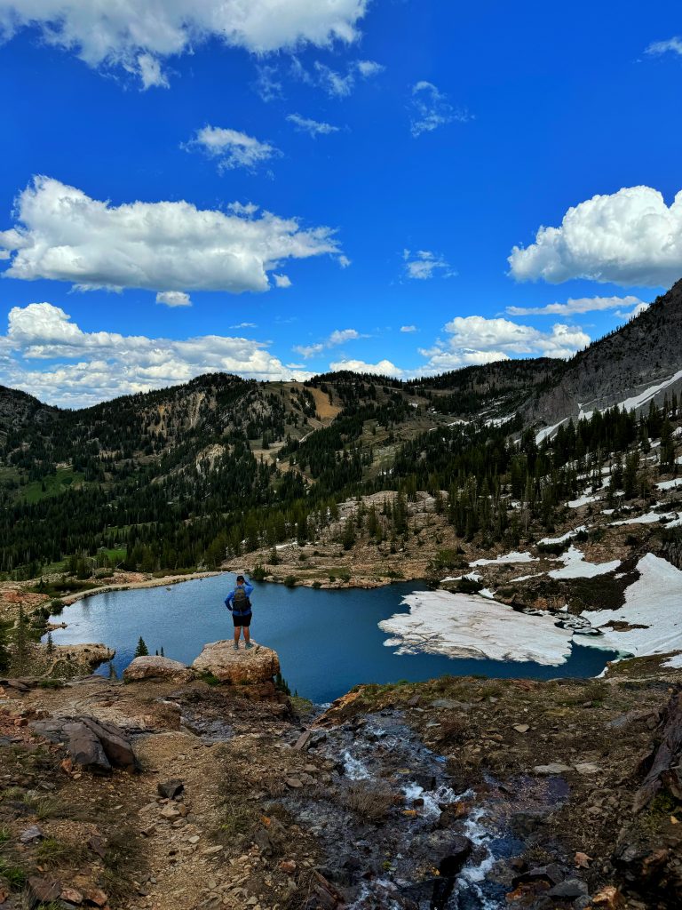 Man stands on a rock with a beautiful blue lake in the background partially frozen and surrounded by mountains