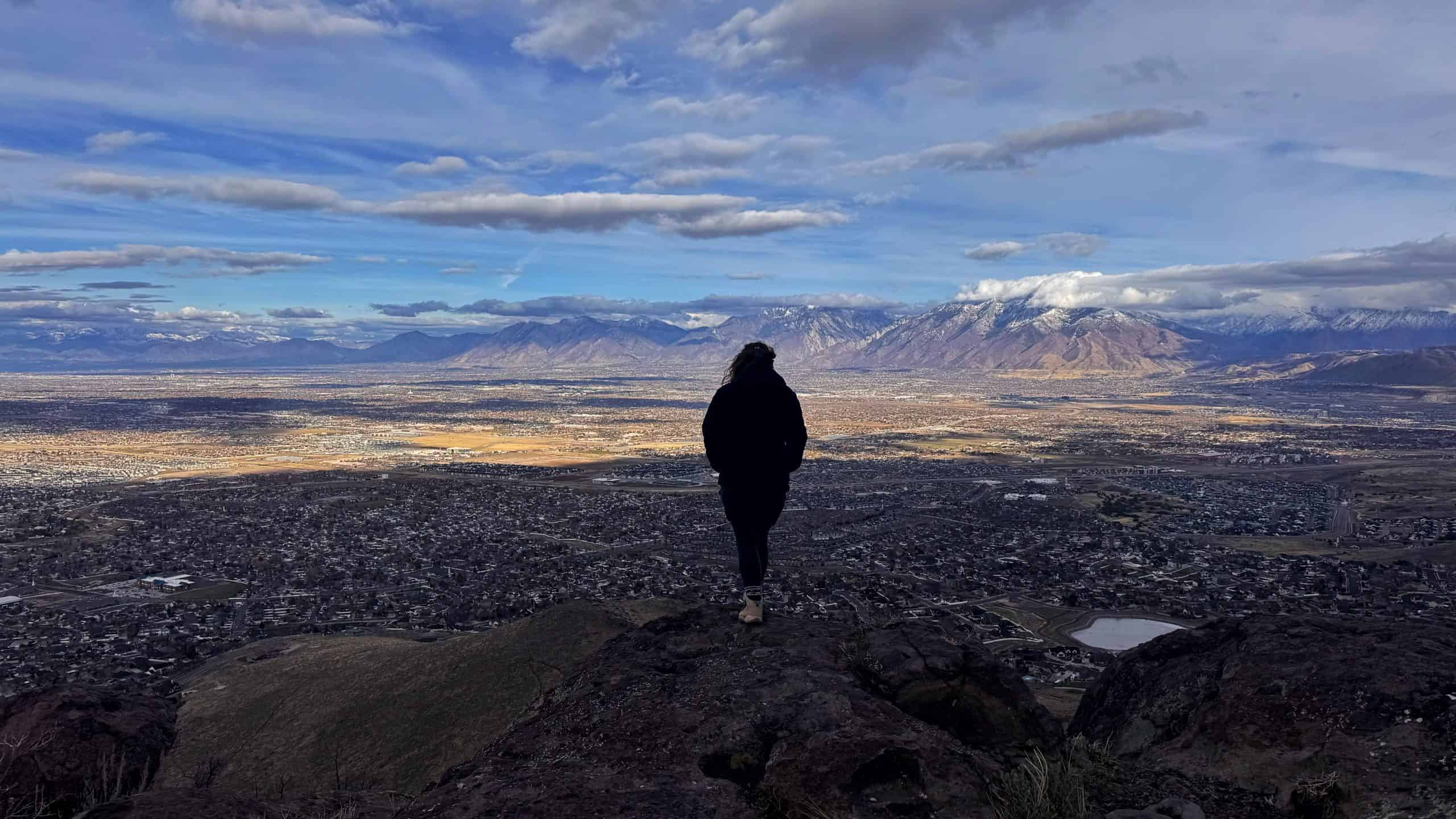Woman standing on a rock, overlooking the vast Salt Lake Valley, with distant mountain peaks. 