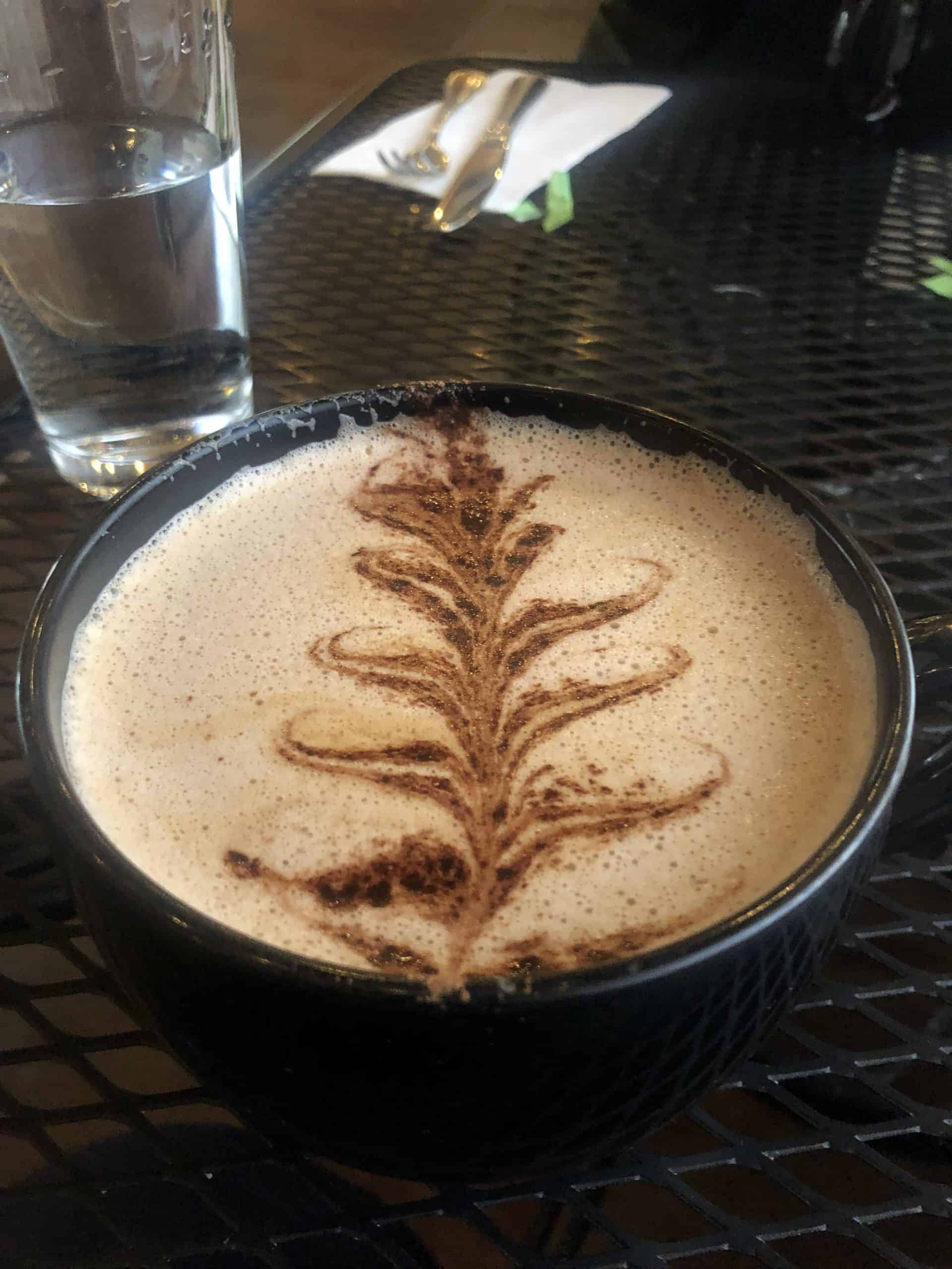 A cappuccino in a dark cup topped with detailed leaf latte art, sitting on a café table with a glass of water nearby.
