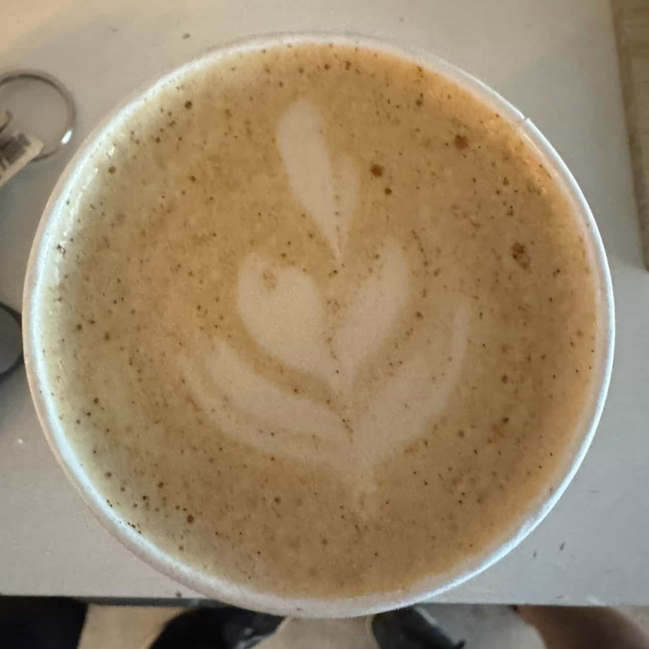 An overhead view of a latte in a white cup with a simple leaf-shaped latte art on a light-colored table.
