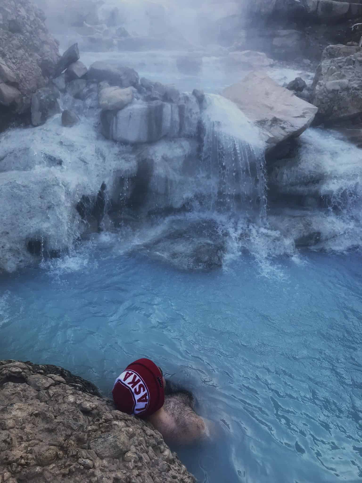 A man sitting at the edge of a steaming turquoise hot spring, wearing a red beanie, with mist rising over rocks and waterfalls in the background.