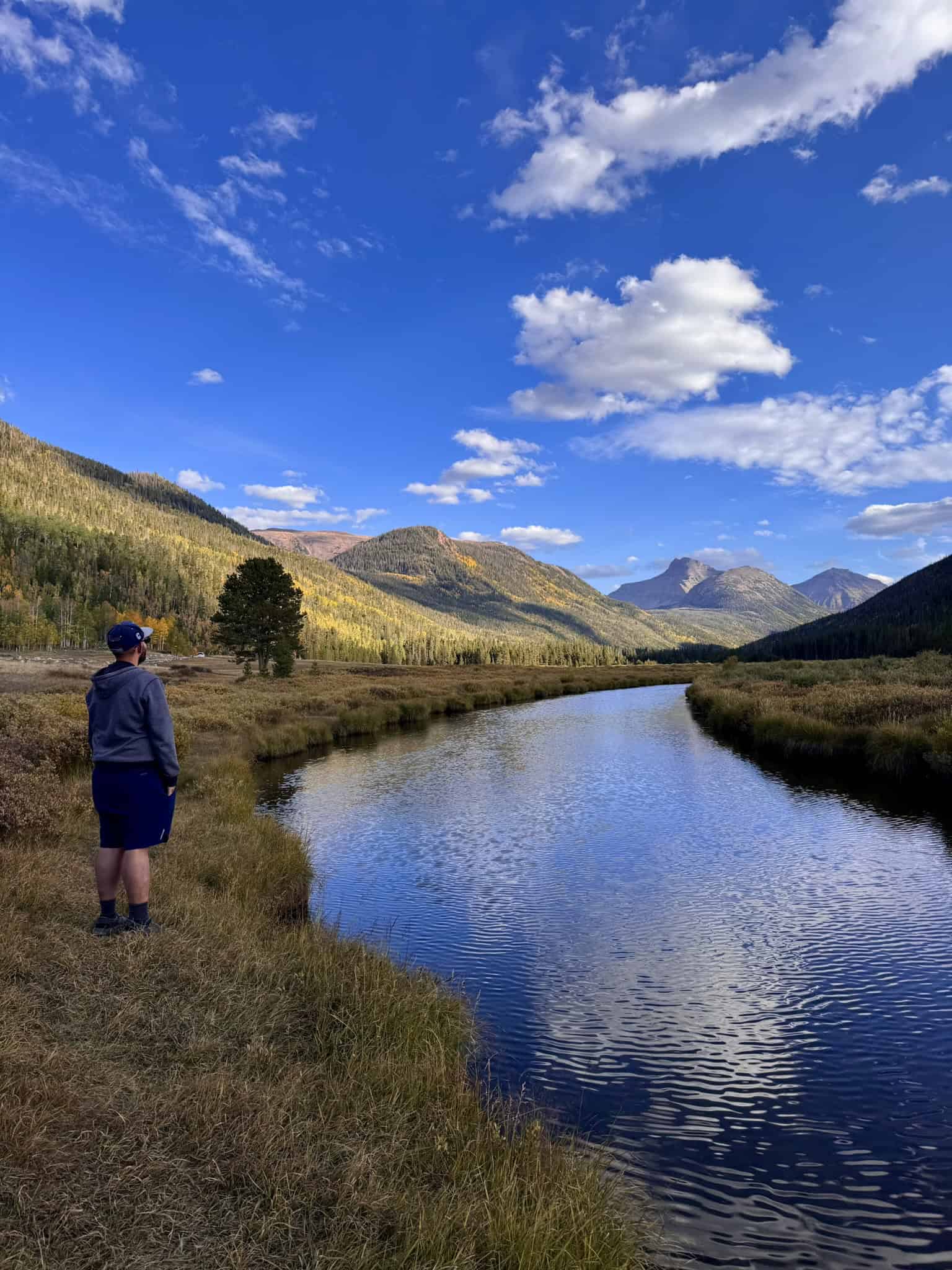Peaceful landscape with a wide calm river, leading to distant rugged mountains, blue skies with a mix of white clouds, and a man standing on the right corner gazing. into the distance. 