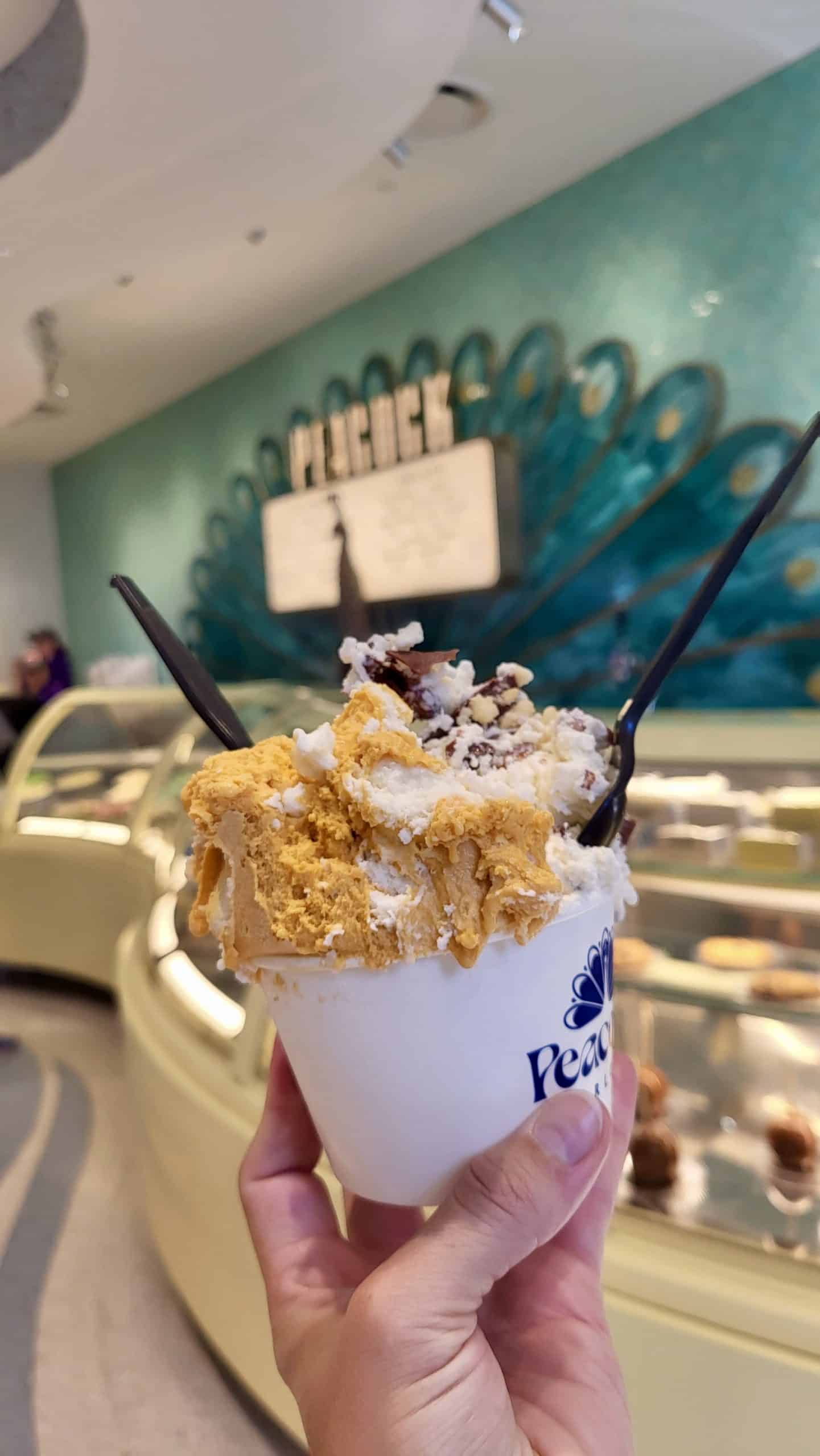 An over-filled gelato cup with two spoons sticking out, served inside a gelato shop.