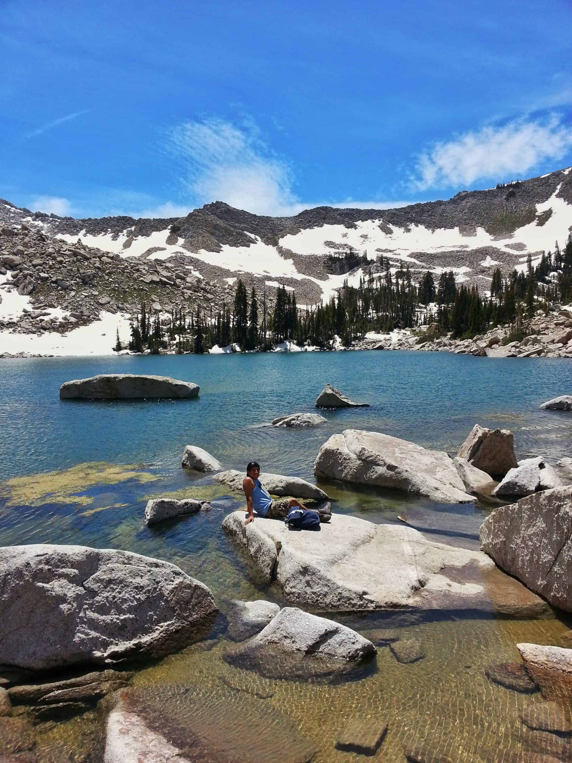 Hiker resting on shoreline rocks beside Upper Red Pine Lake, surrounded by snowy peaks and clear blue water on one of the most scenic hikes near Salt Lake City.