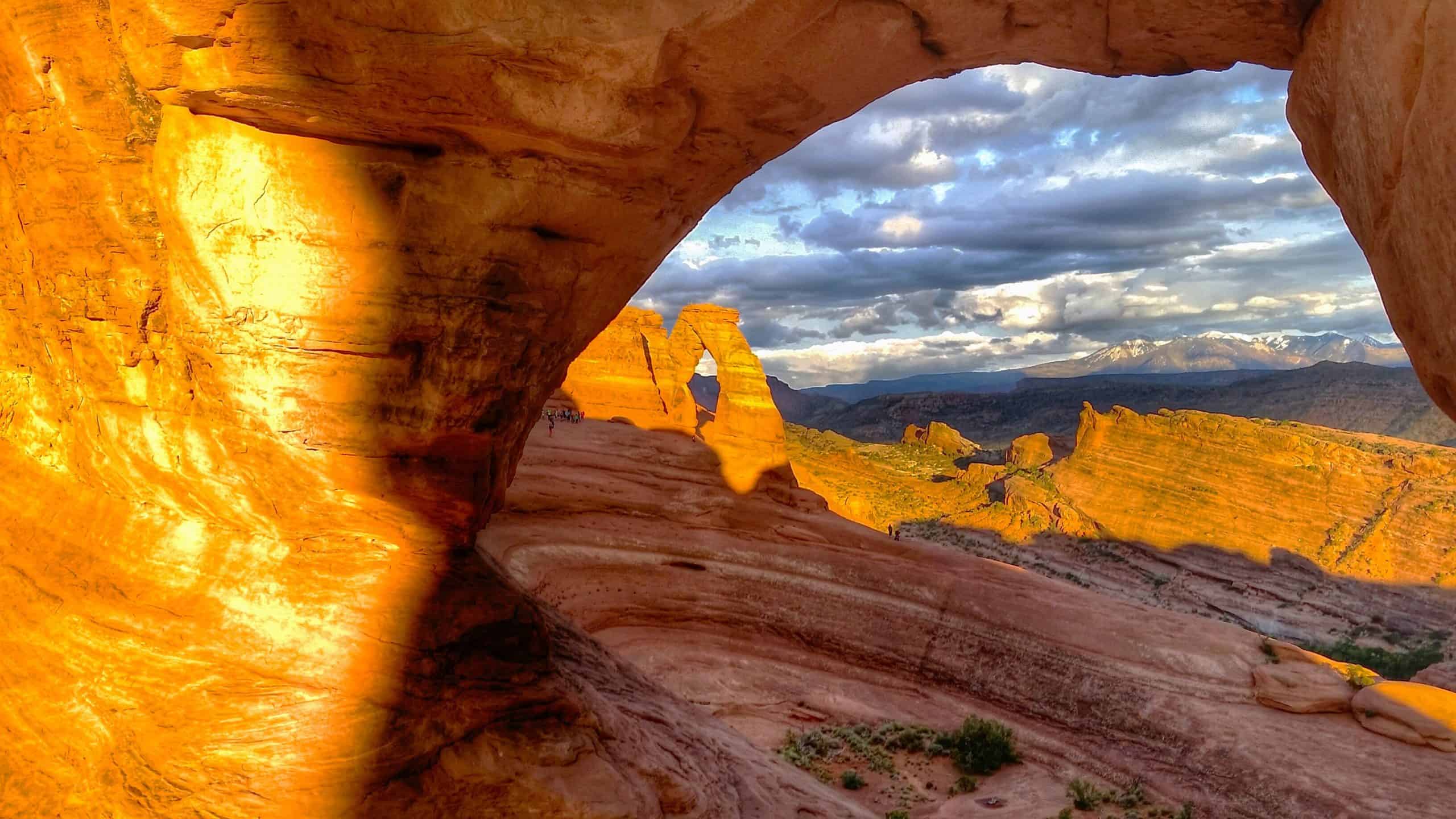 Delicate Arch in Arches National Park, Utah glowing at sunset, framed by red sandstone with distant mountains and clouds.