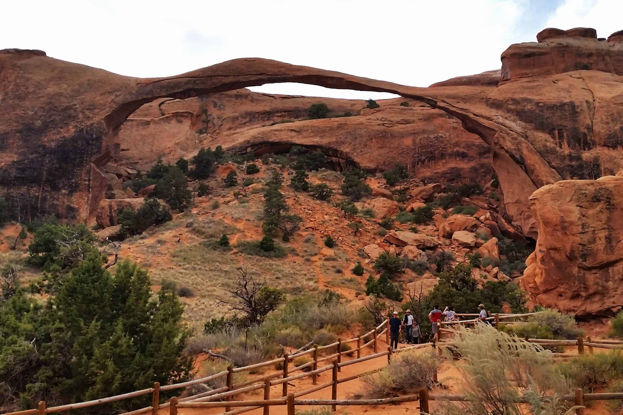 Landscape Arch spanning a desert trail with red rock cliffs and hikers below in Arches National Park, Utah