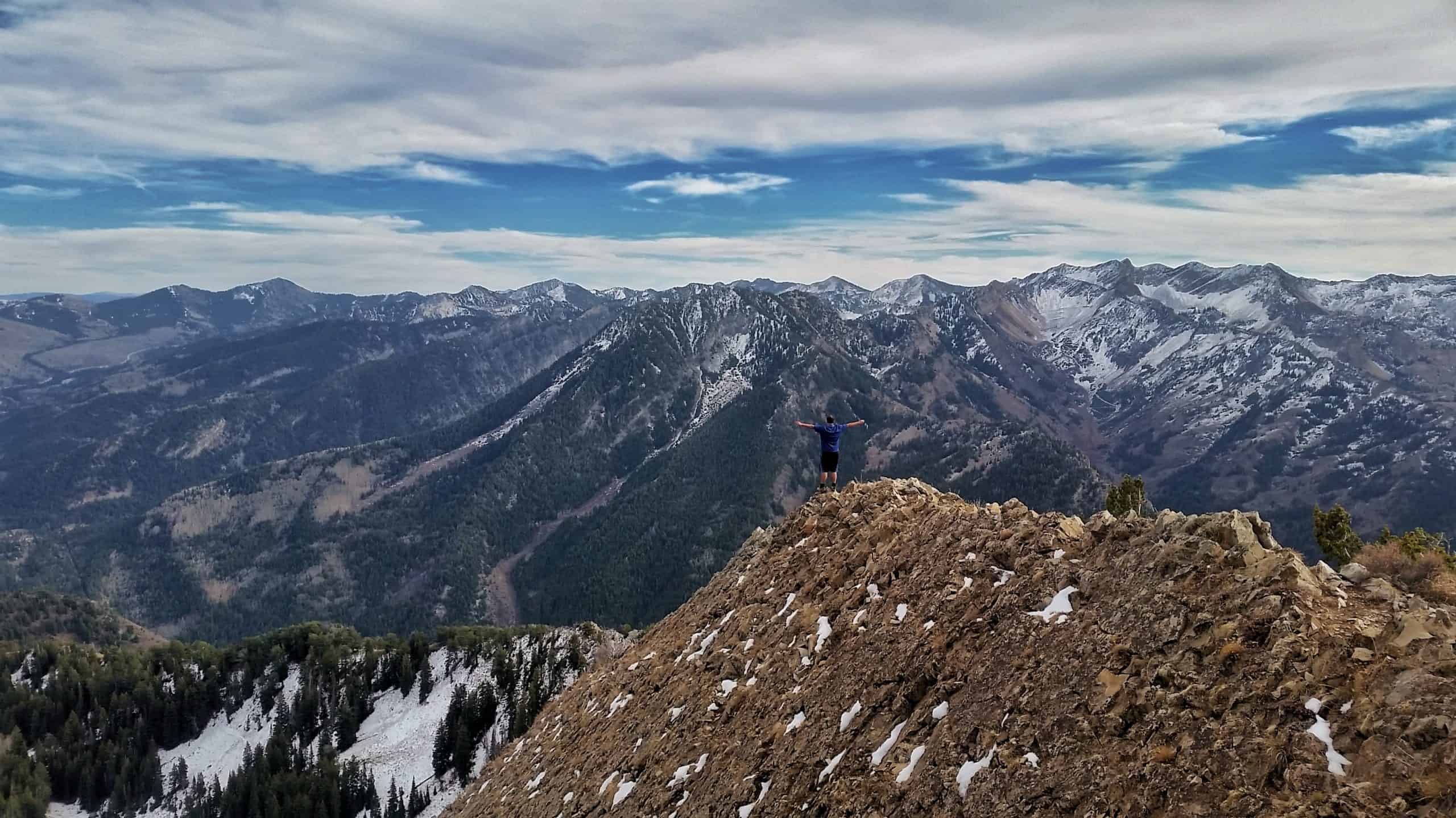 Hiker standing on Mount Raymond summit overlooking rugged, snow-dusted mountains in Big Cottonwood Canyon.