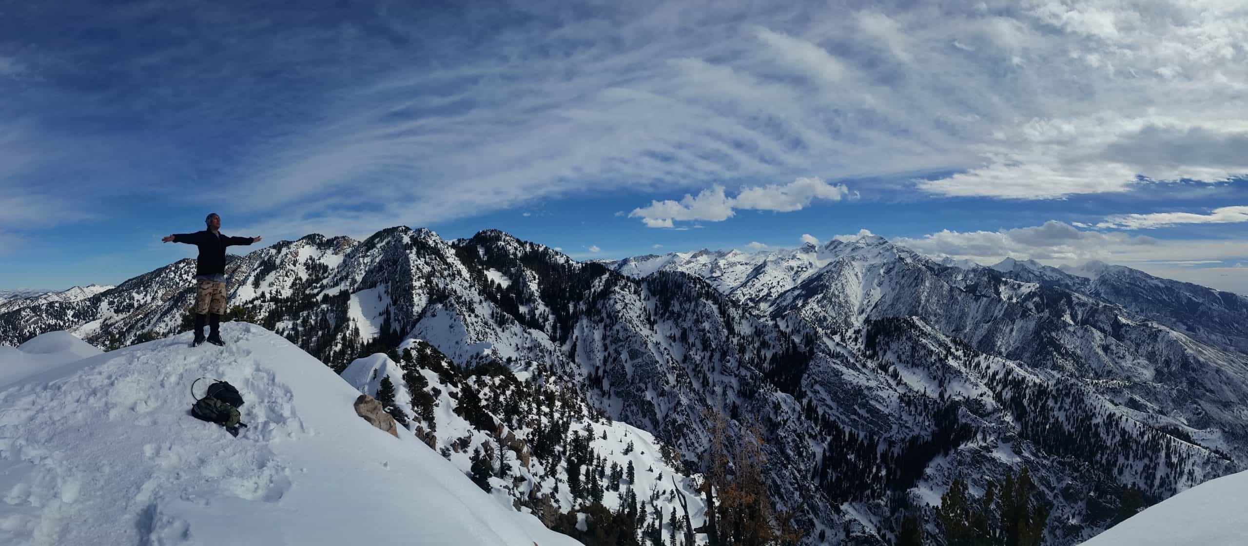 Hiker stands with arm's stretched out on the summit of Mount Olympus near Salt Lake City, with snow blanketing all the nearby mountains