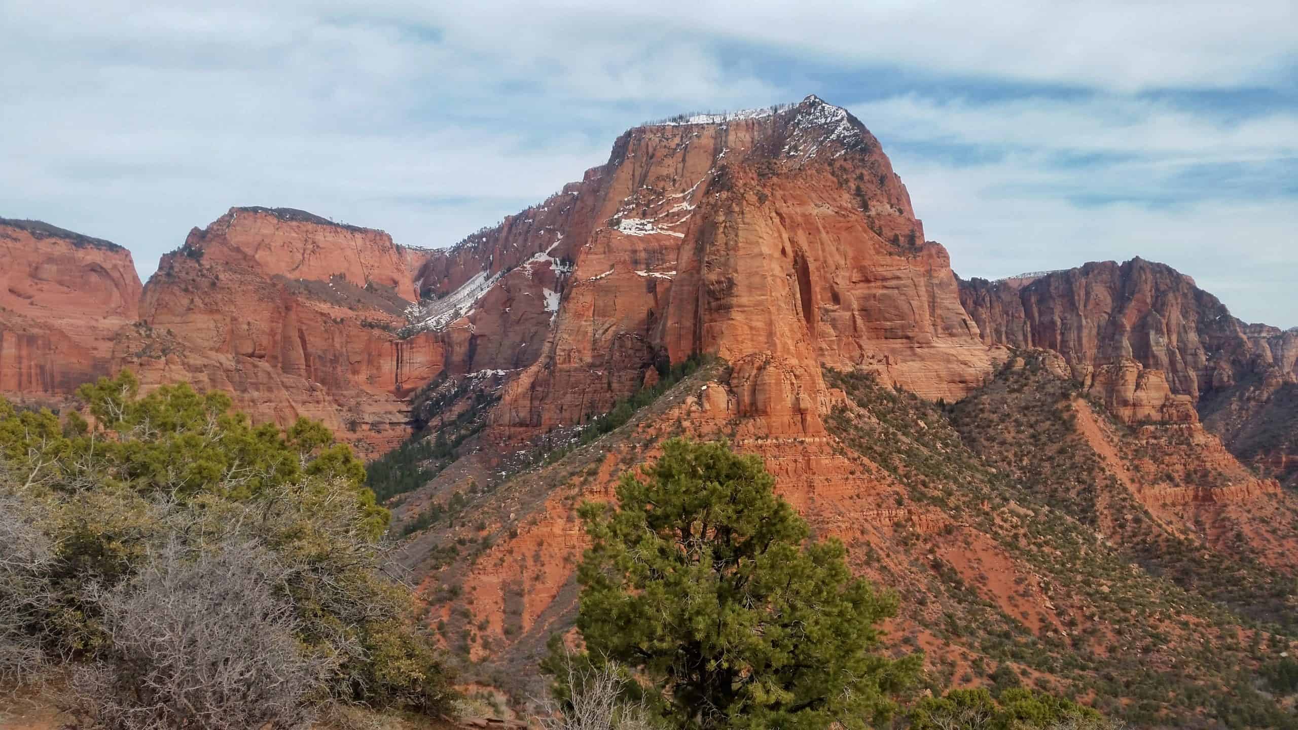 Tall sandstone walls with a dusting of snow, towering against a partly cloudy sky in Kolob Canyon, Utah.