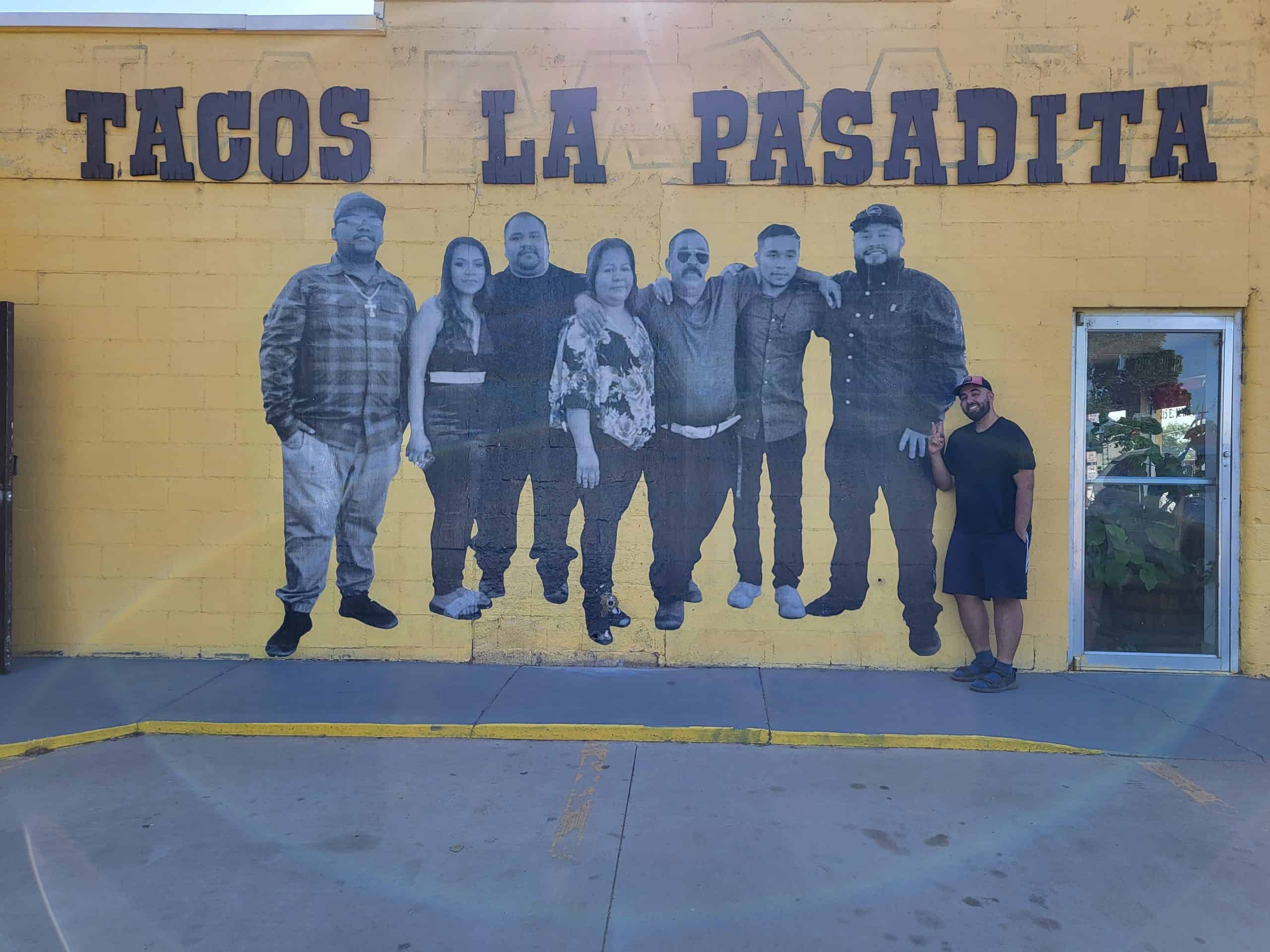 Man standing next to a yellow brick wall with a photo of a family of smiling people with the words "Tacos La Pasadita" written above. 