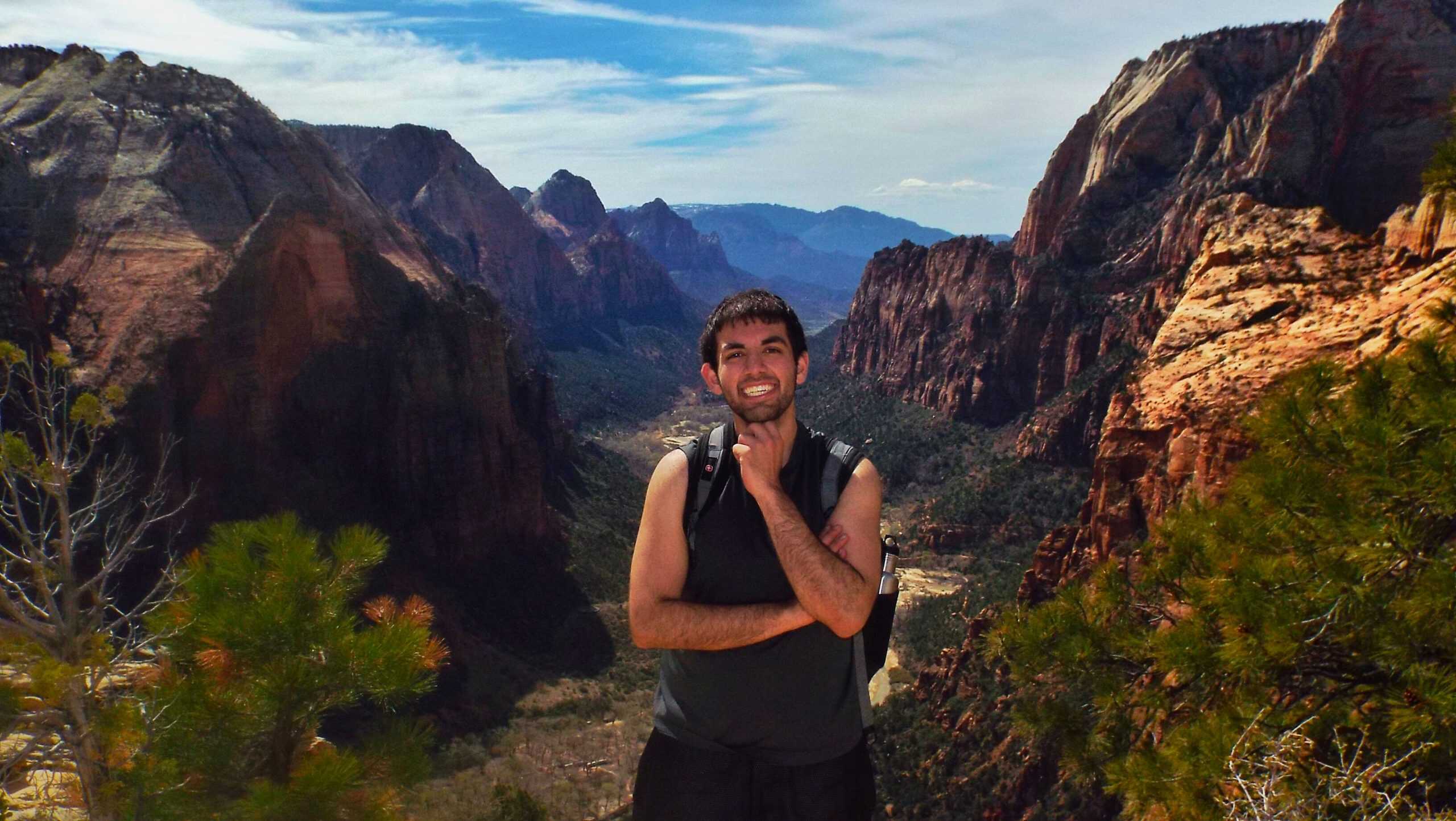 A hiker standing on the summit of Angel's Landing, a thrilling hike in Utah, smiling at the camera while wearing a backpack on a narrow ridge surrounded by dramatic red rock cliffs and vast canyon views below.

