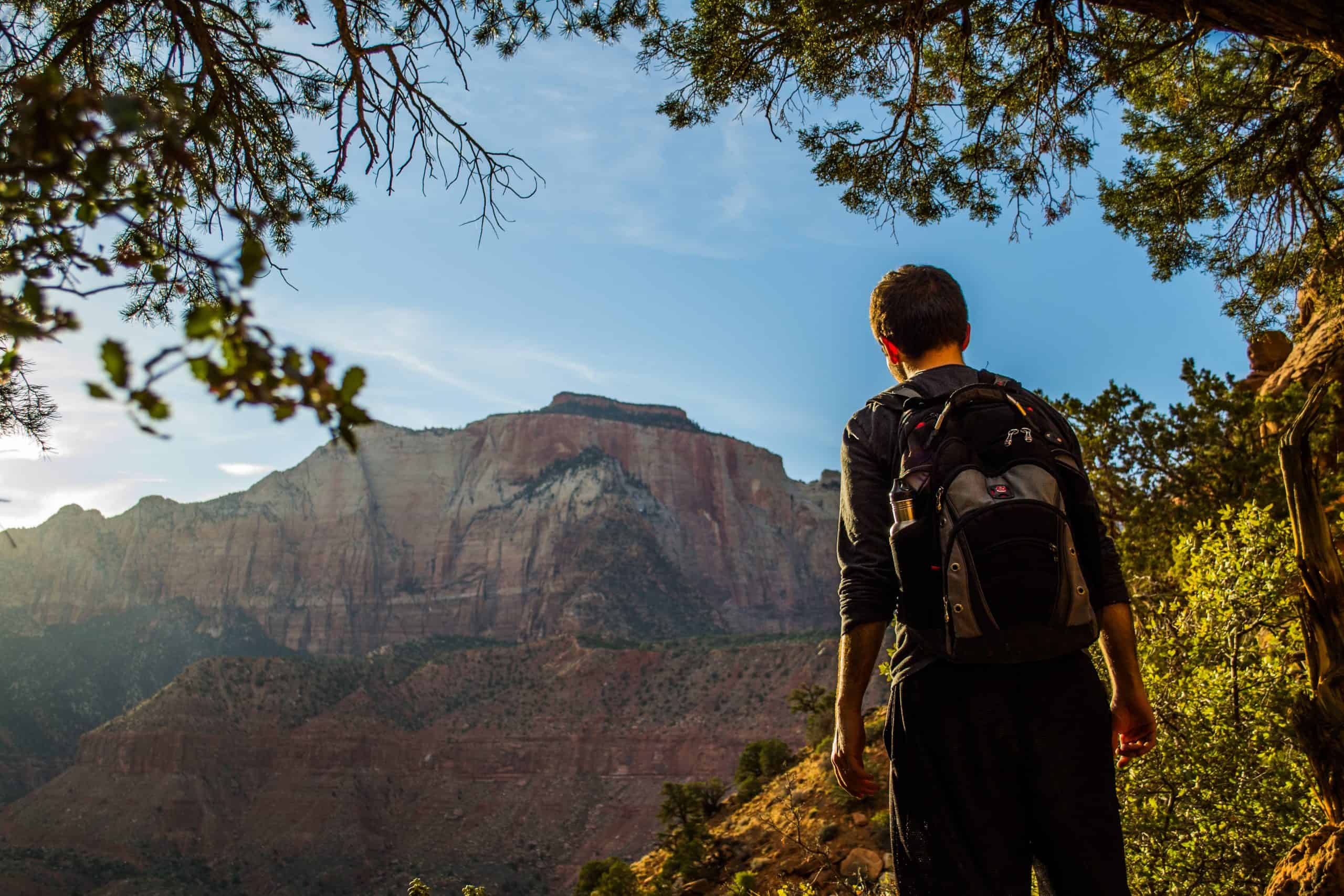 Man standing looking away from the camera with a distant towering sandstone cliff, framed by trees. 