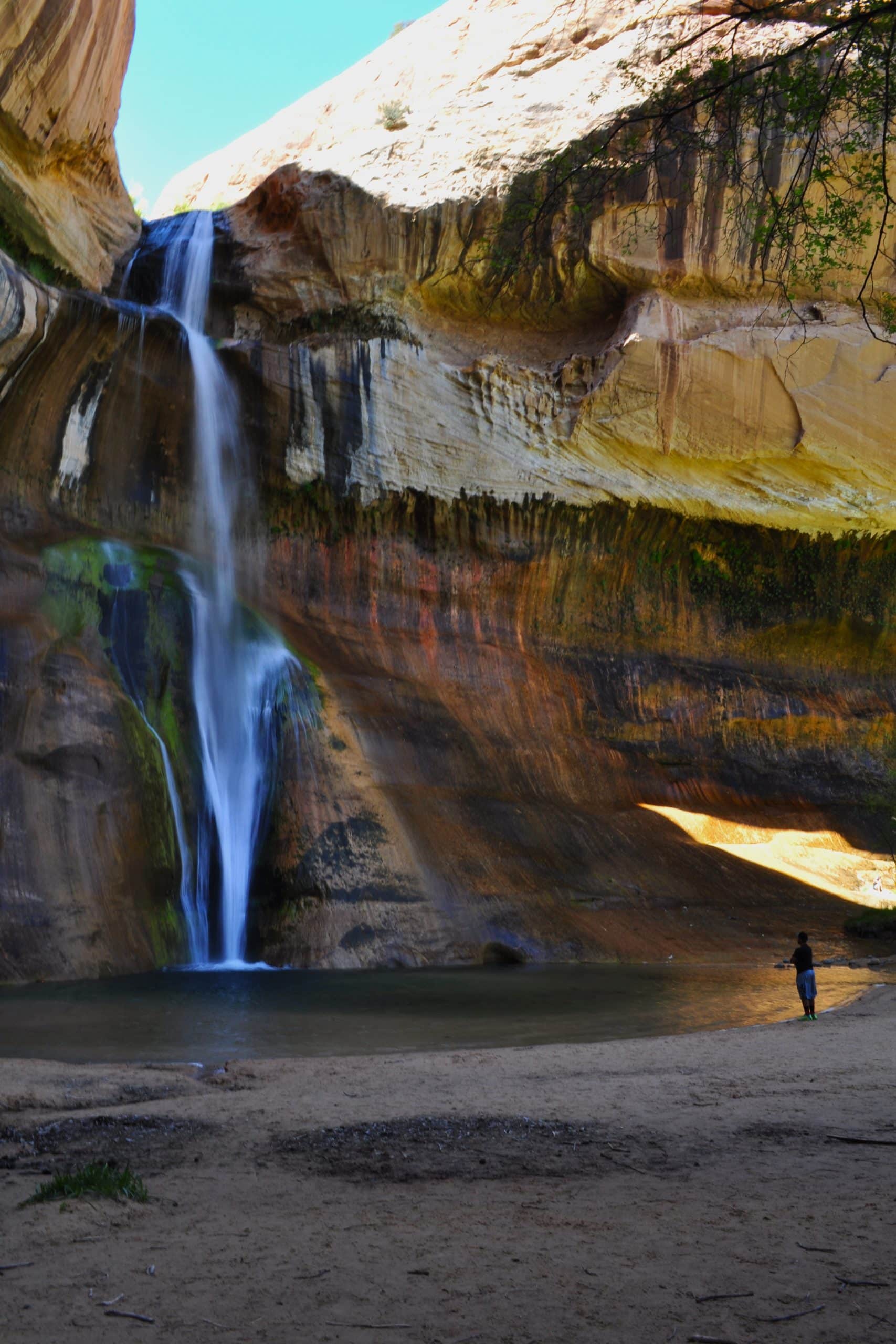 A person enjoying one of the best hikes in Utah, gazing up at a majestic waterfall, watching it cascades down a sandstone cliff face into a pool below