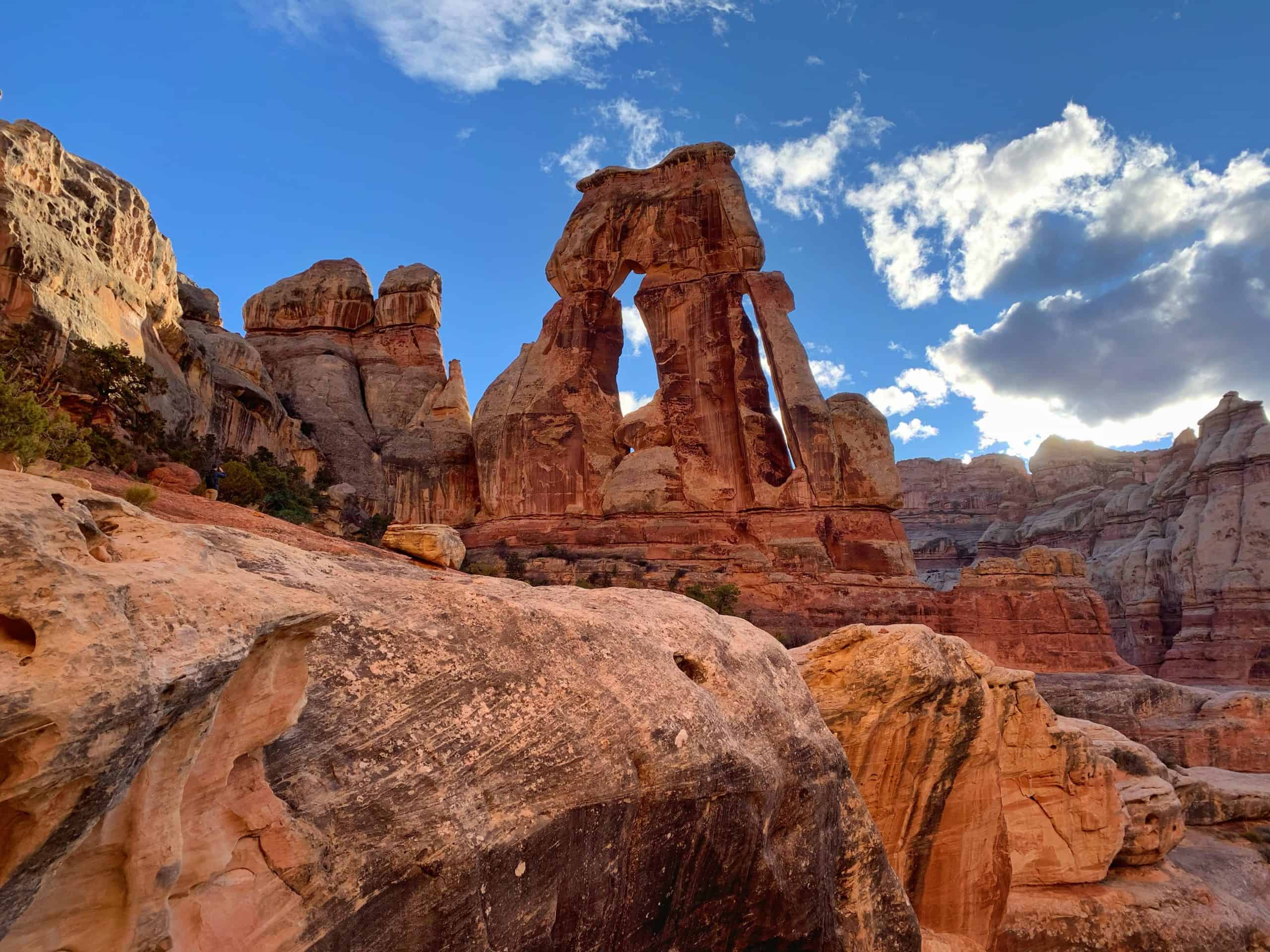 A massive red sandstone arch formation stands tall against a blue sky with scattered clouds.

