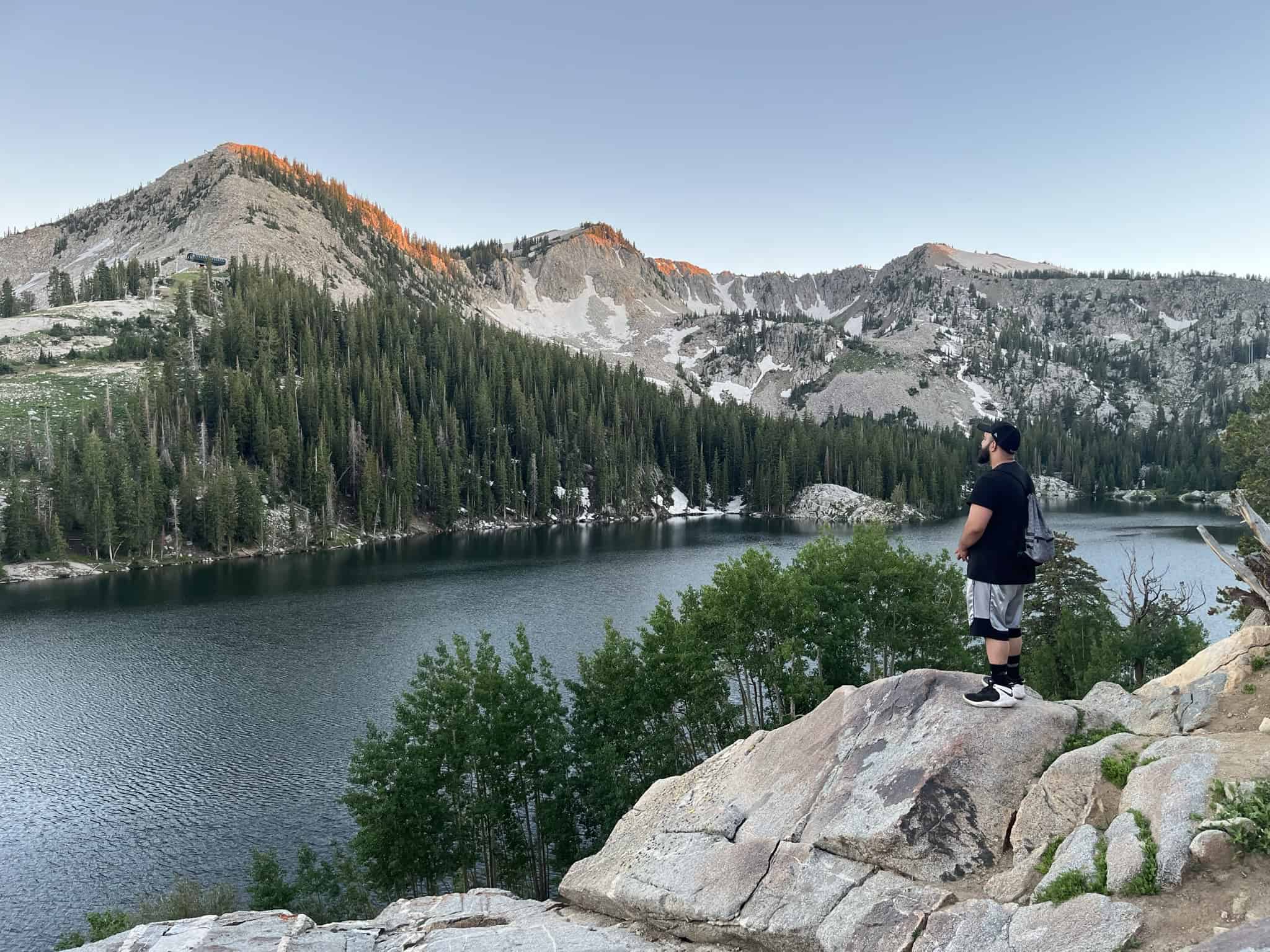 Hiker standing on a rocky overlook above Twin Lakes with evergreen trees and rugged peaks in the background.