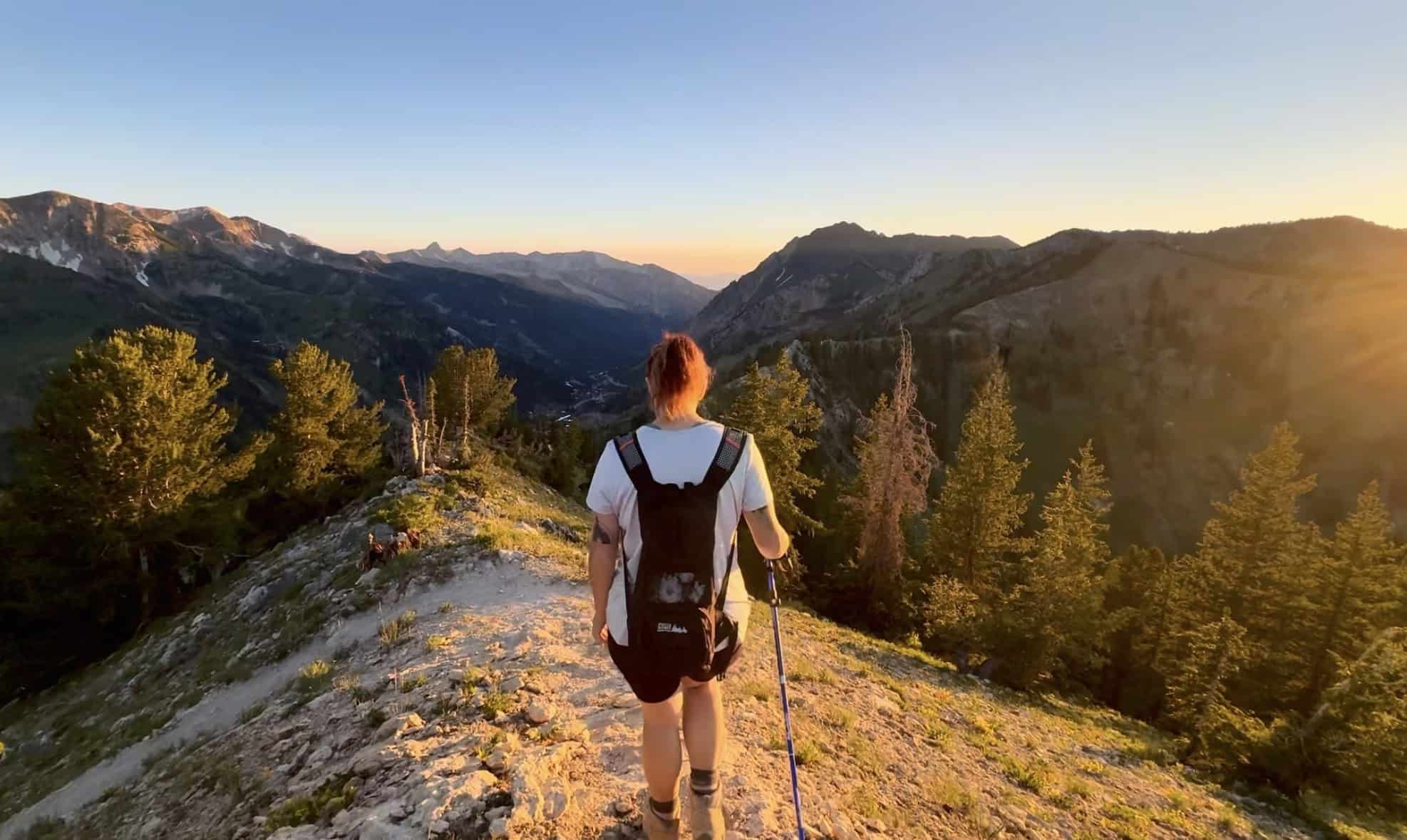 Women hiking down a mountain ridge, surrounded by pine trees and distant mountain peaks with golden hour lighting emerging from the right