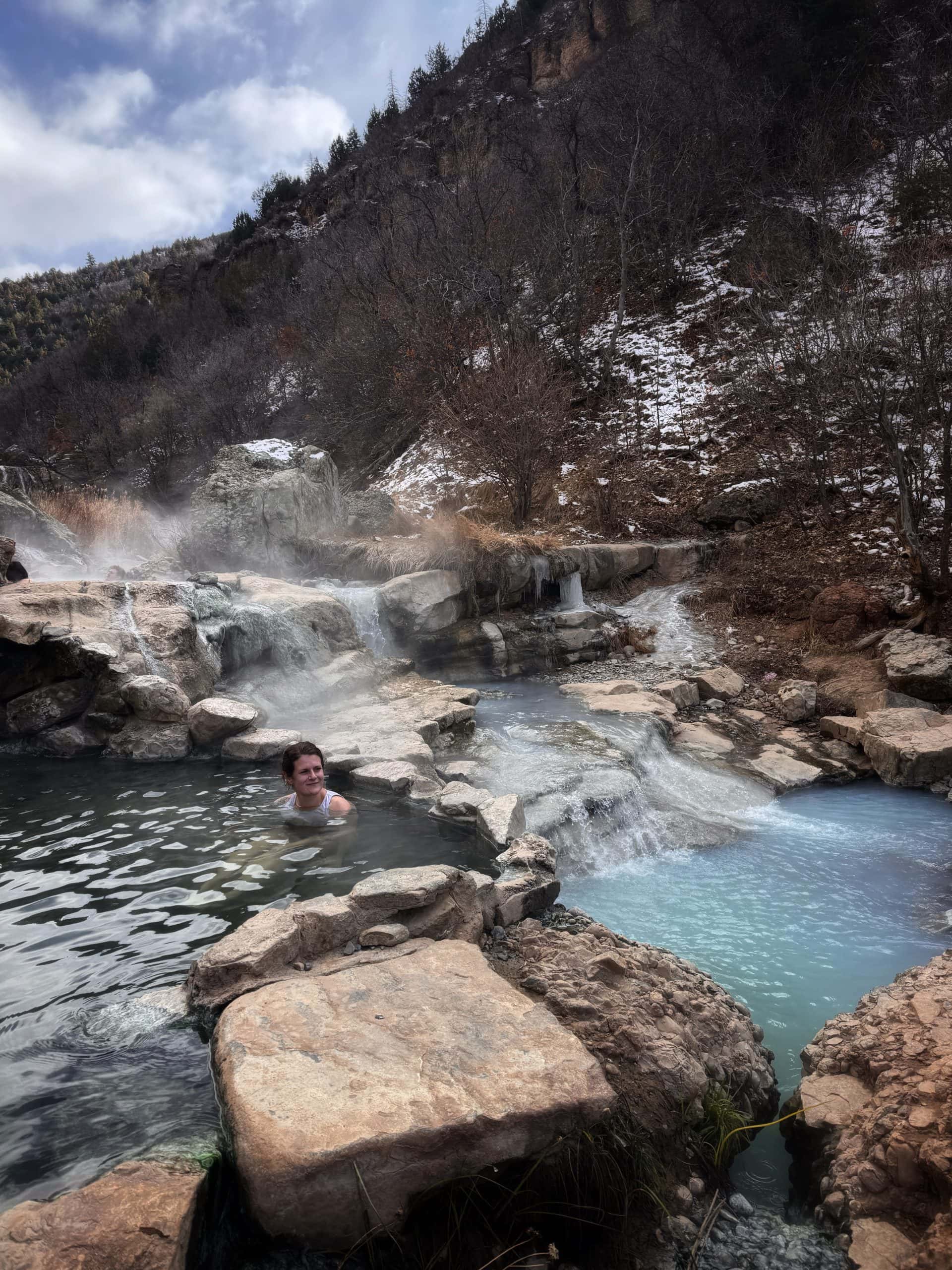 A woman sits in a steaming turquoise hot spring surrounded by boulders, with a forested backdrop dusted in snow 