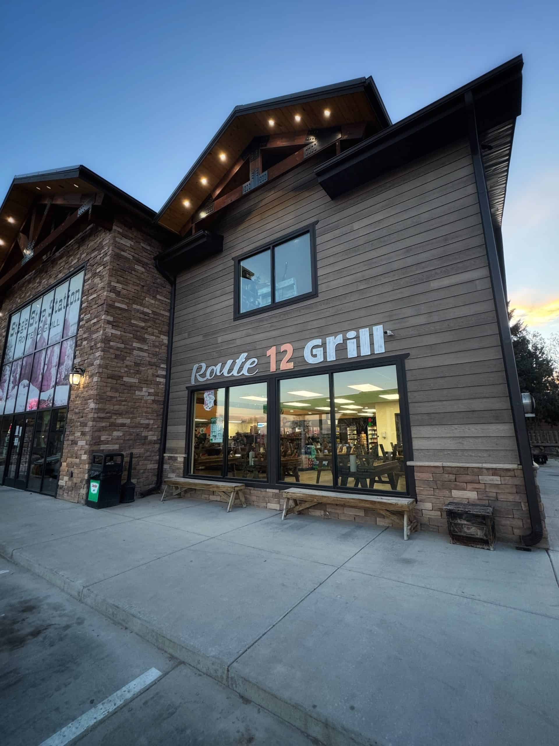 A modern, two-story building with wood and stone siding, featuring large windows and an illuminated “Route 12 Grill” near Bryce Canyon National Park, Utah. 


