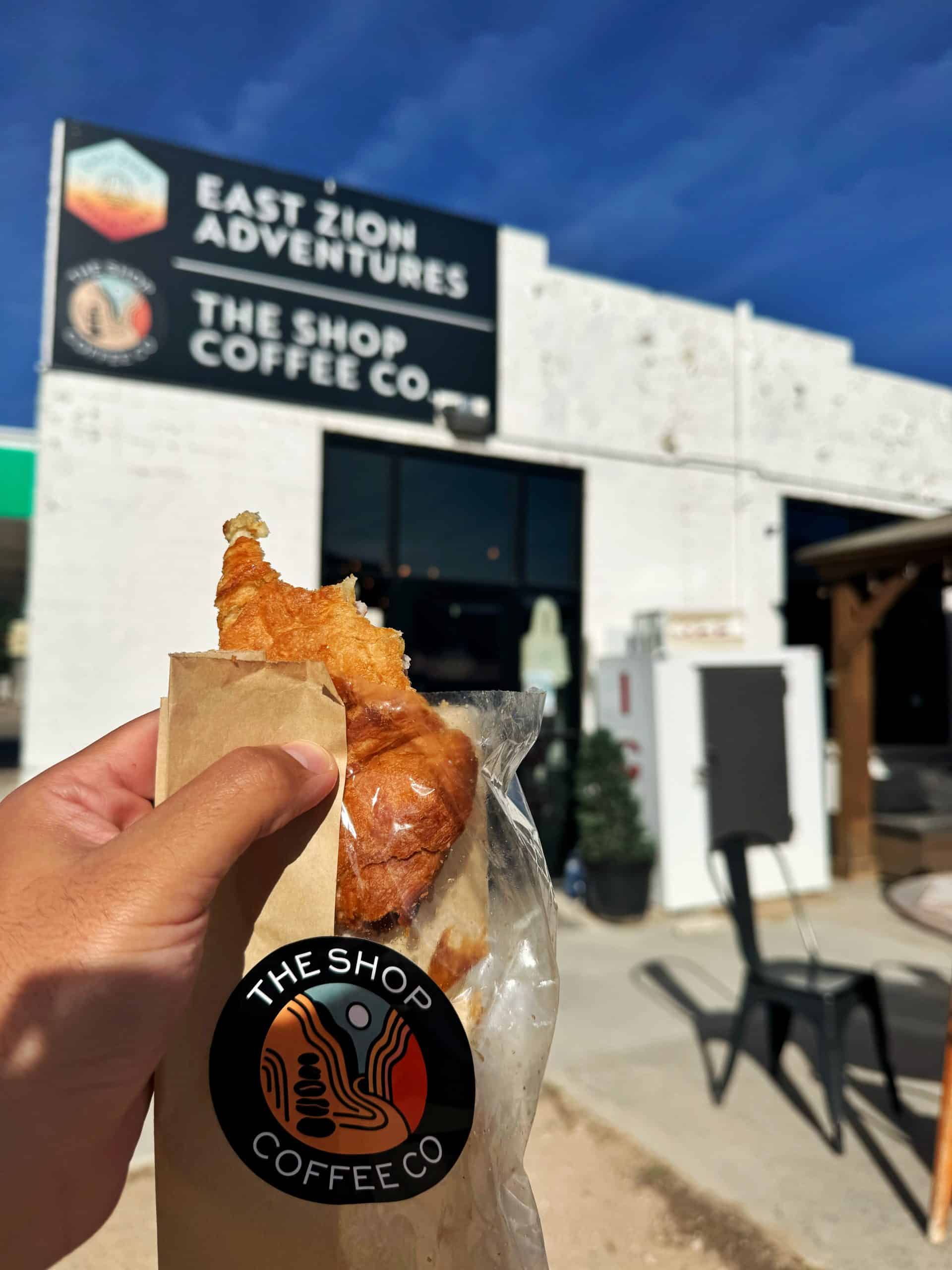 A hand holds a croissant from The Shop Coffee Co. with its sign visible in the background during a stop on the Utah mighty 5 national park road itinerary.

