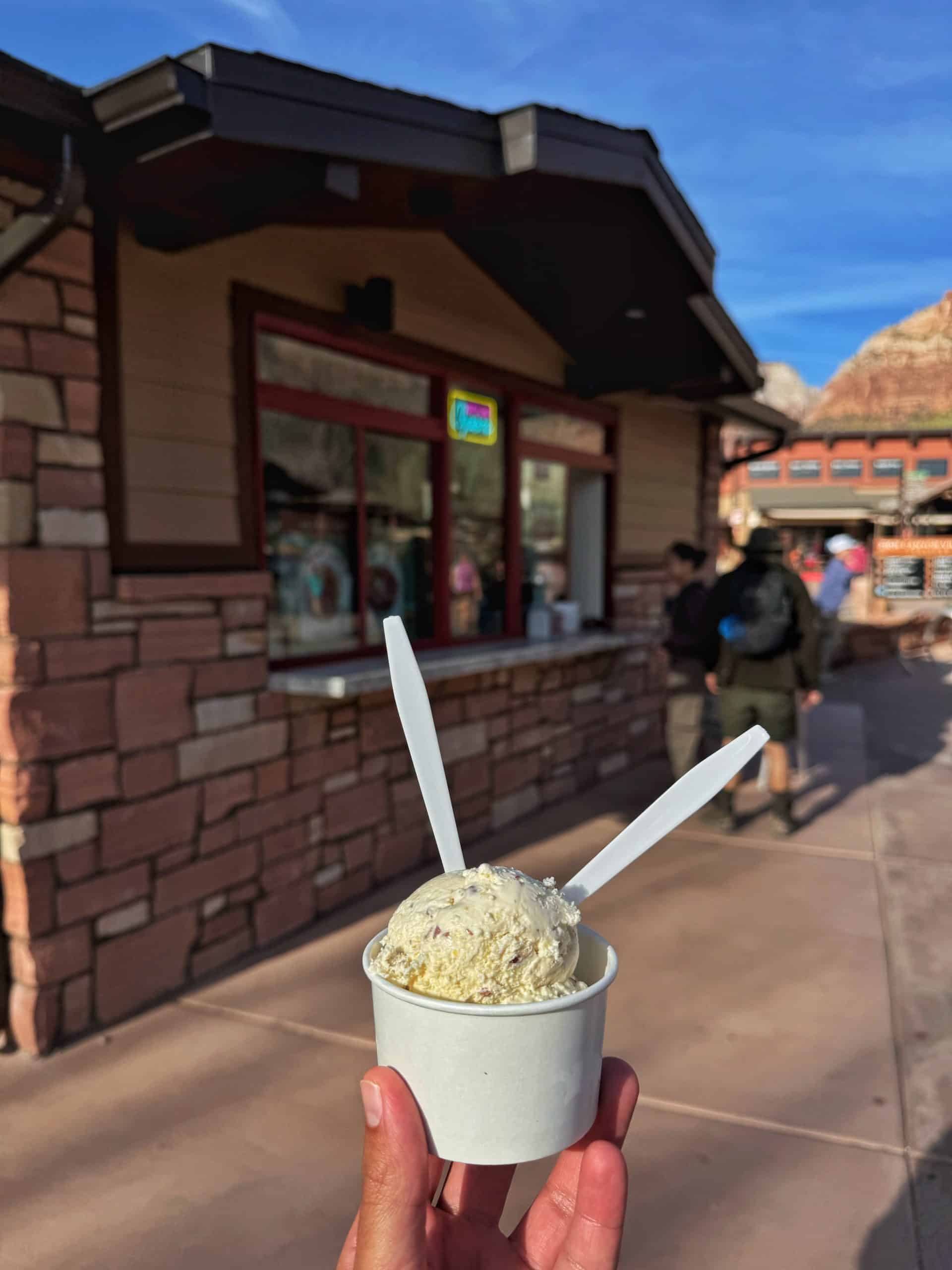 A hand holds a cup of salted caramel Gelato from Desert Ice at a stone building in Zion National Park,
