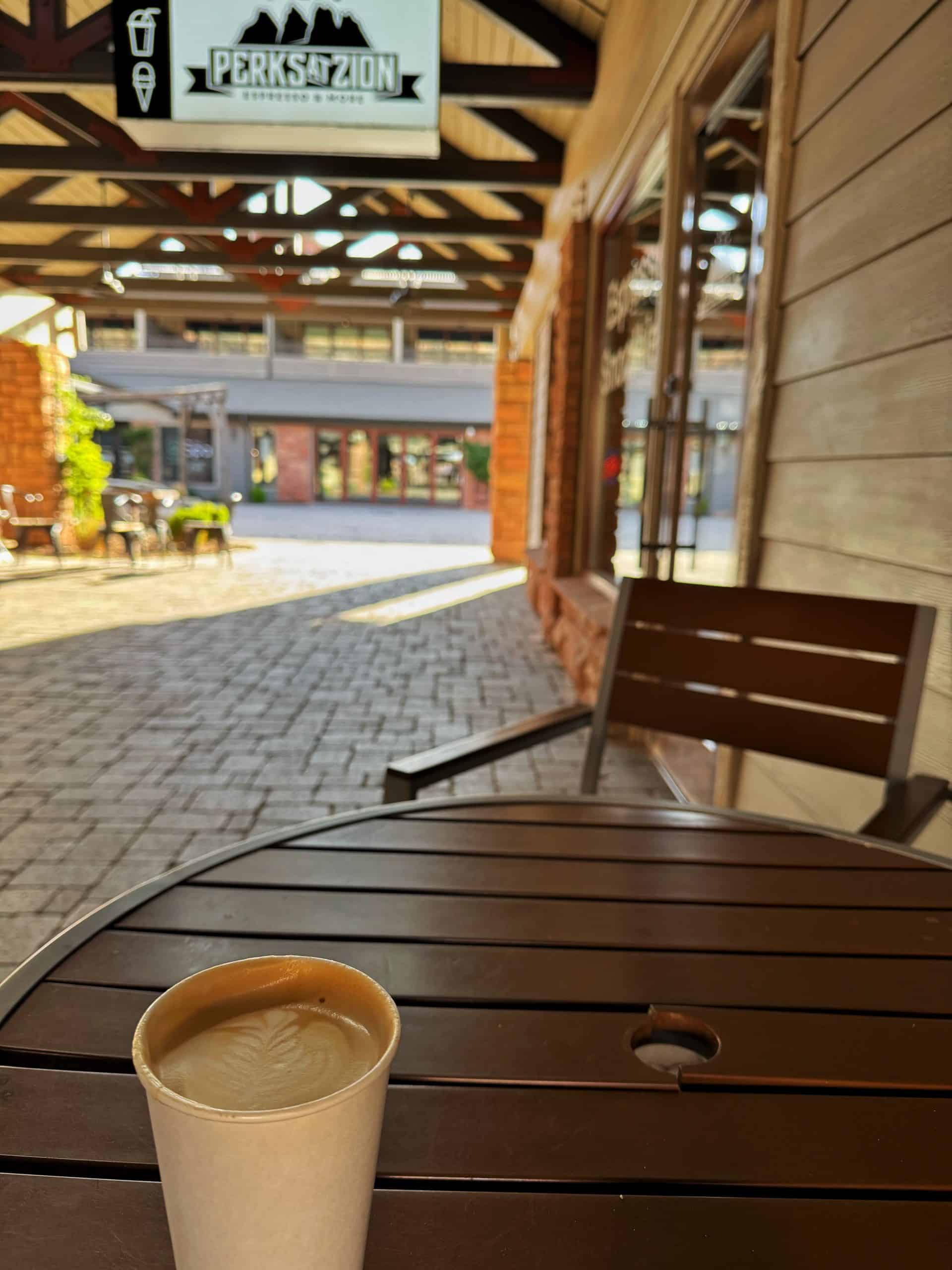 A coffee cup with latte art sits on a wooden table outside of Perks At Zion under a shaded walkway


