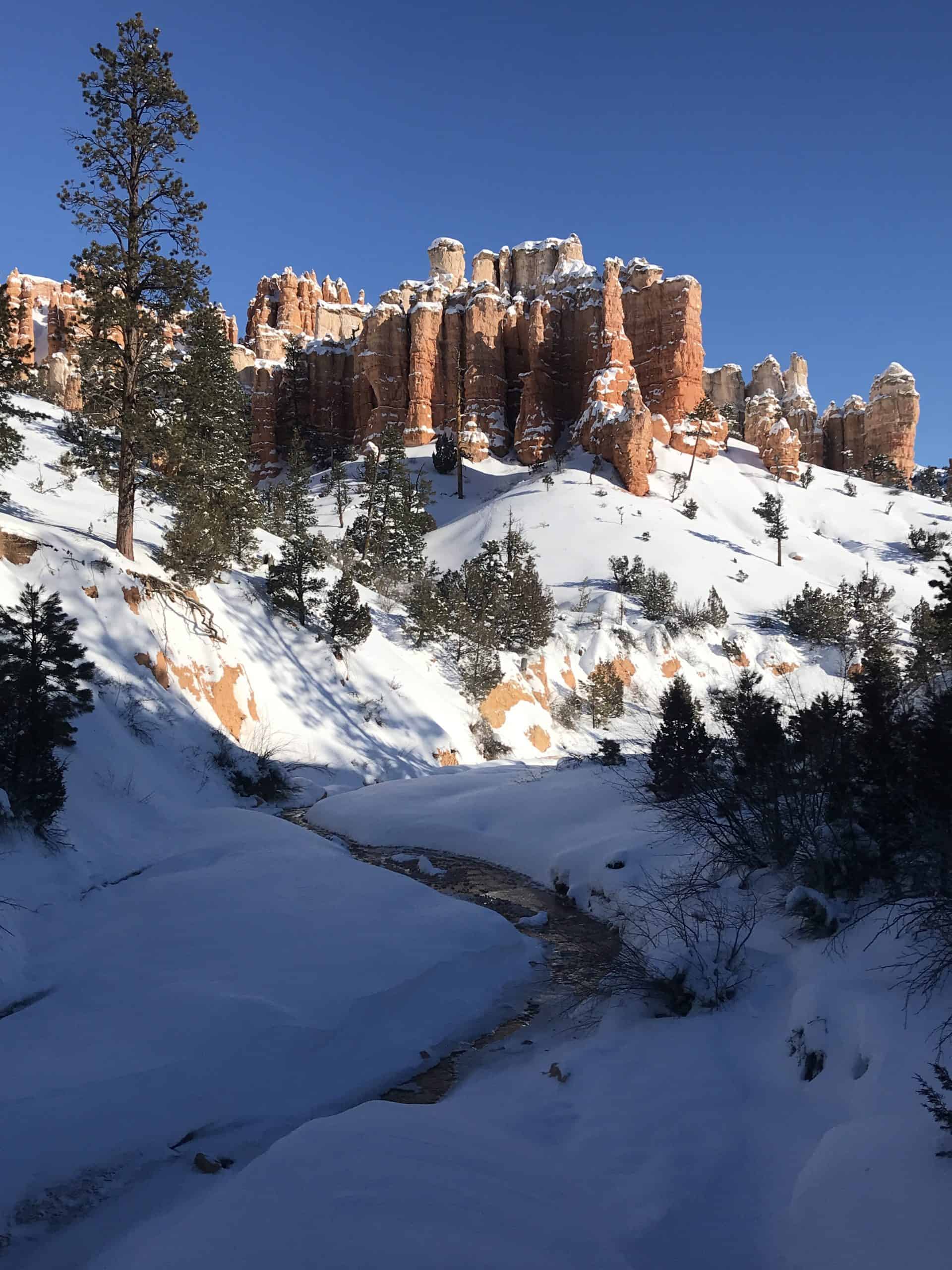 Snowy river leading to a stunning view of tall orange hoods and evergreen trees under a deep blue sky on the Mossy Cave Trail in Bryce Canyon National Park, Utah. offering a welcoming stop along the Utah mighty 5 national park road itinerary.
