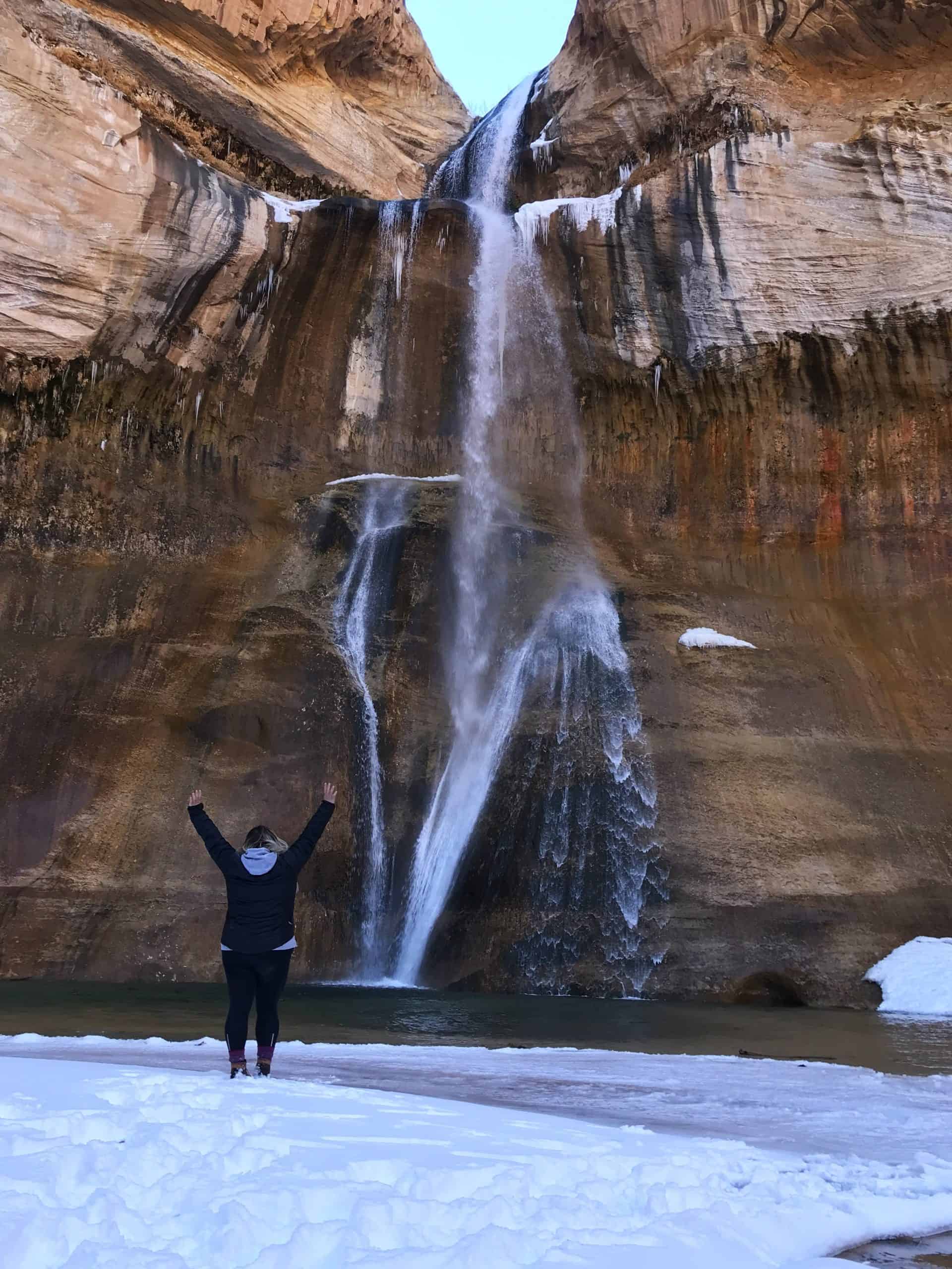 A hiker throws up her arms as she gazes up at a majestic waterfall, watching it cascades down, with snow surrounding the pool below.