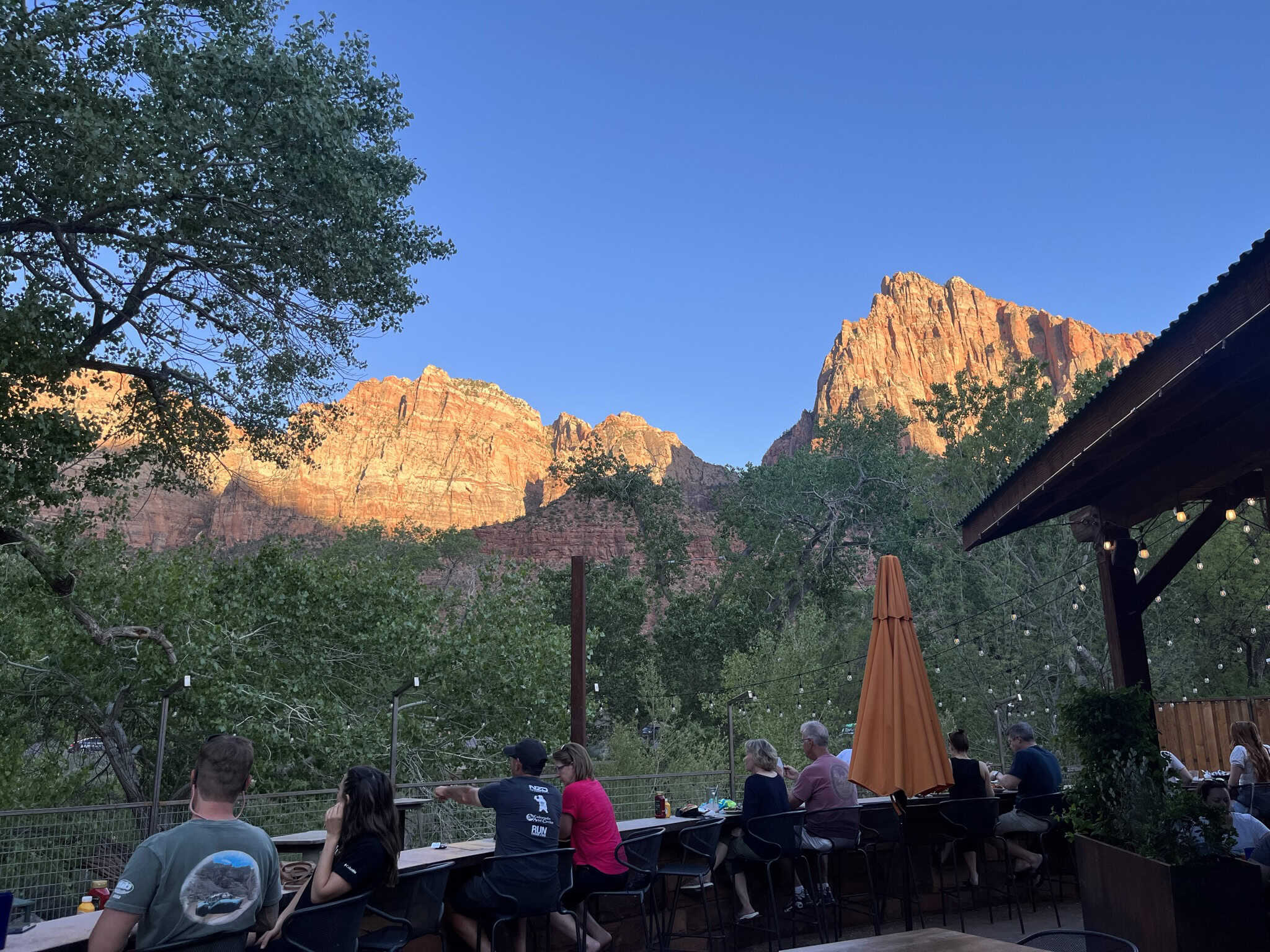 Outdoor Patio with people sitting, set against a background of green trees and towering sandstone cliffs. 