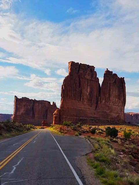 Iconic Courthouse Towers rock formations towering besides the road in Arches National Park, with partly cloudy skies 