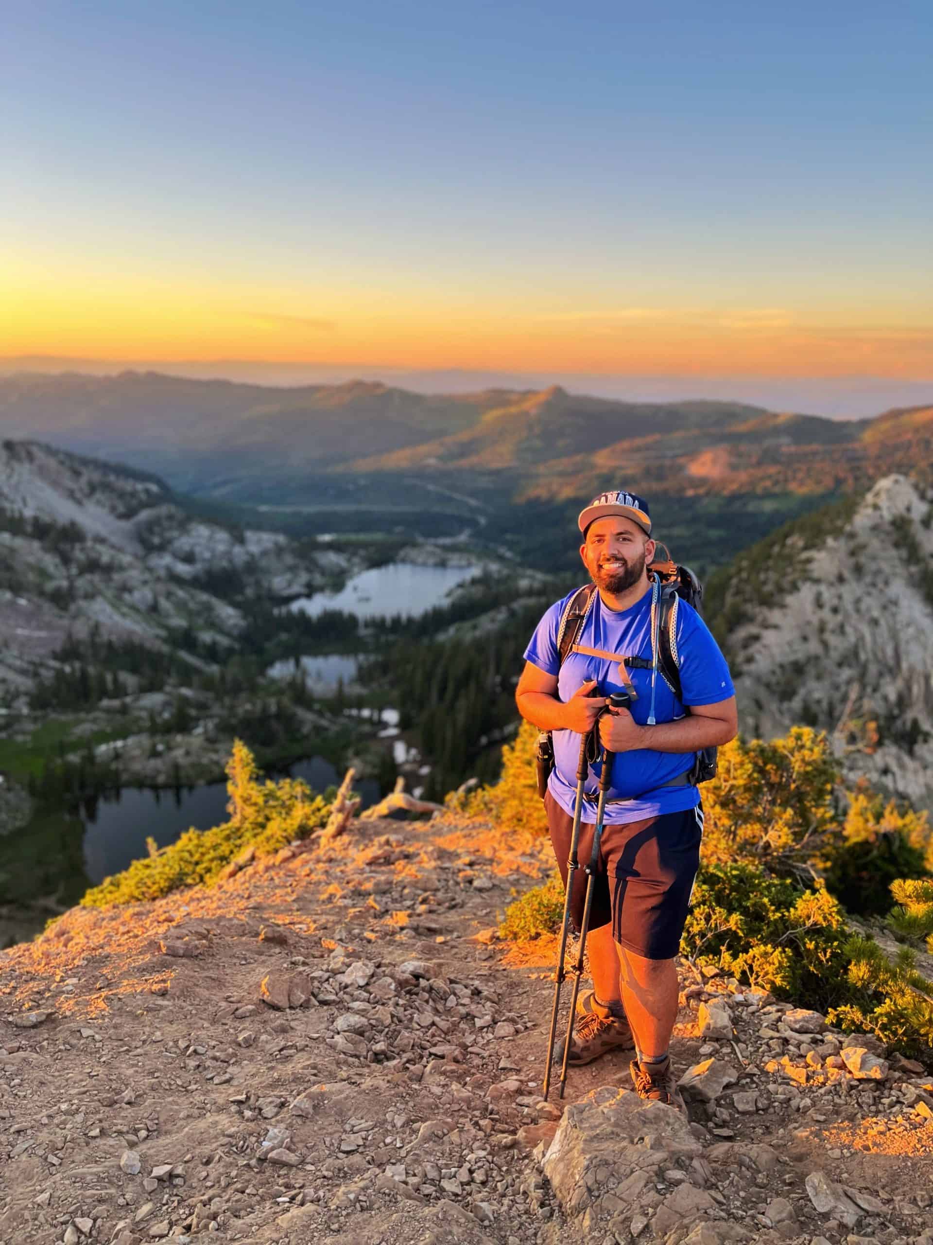 Hiker stands smiling at the camera and holding hiking poles, with the sunset light fading in the background on distant mountains and three scenic lakes in Big Cottonwood Canyon.