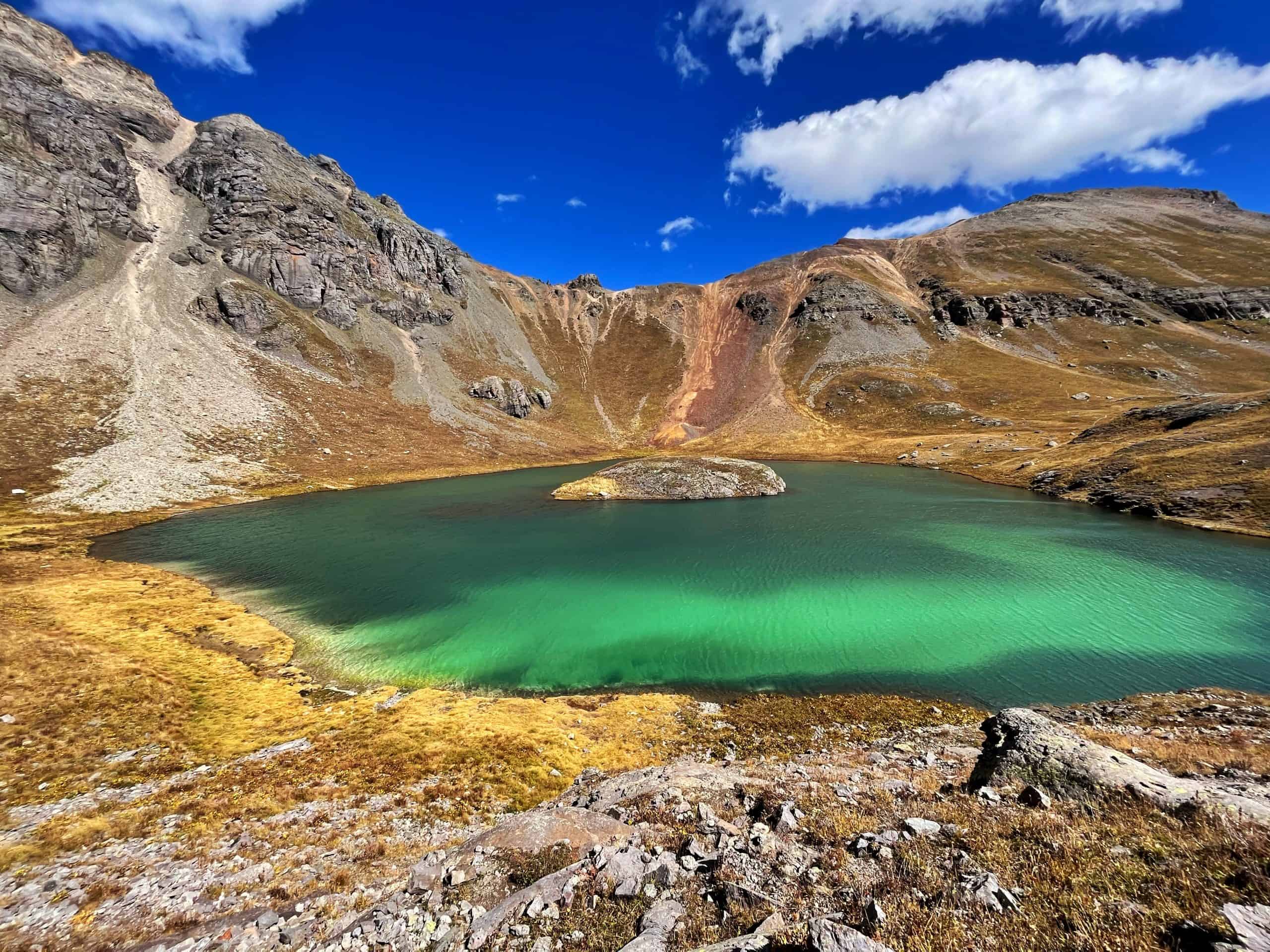 A vibrant emerald lake with a small island nestled in a mountain basin with rocky slopes and autumn-colored vegetation