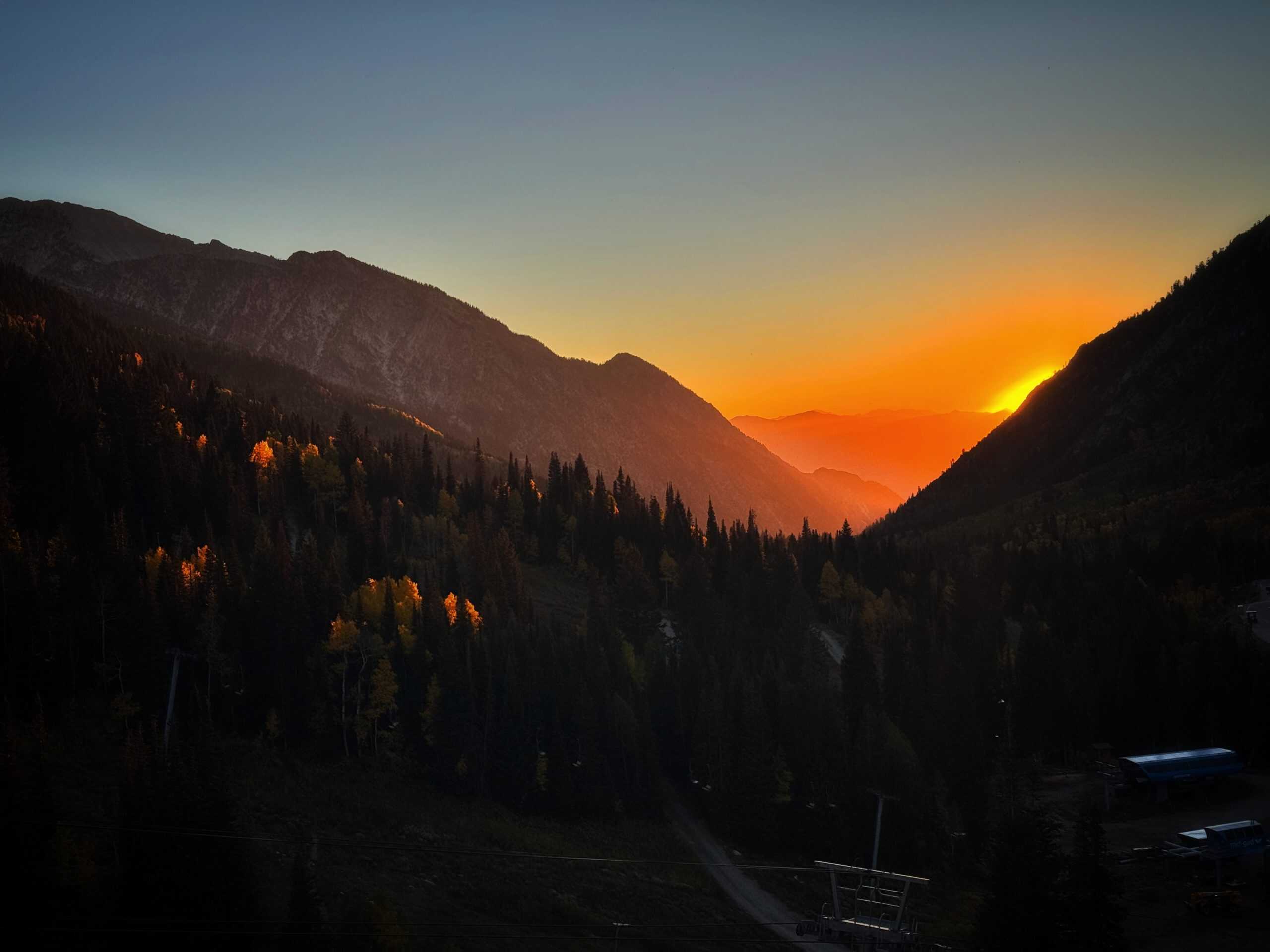 A golden sunset glowing between mountain ridges in Little Cottonwood Canyon, with autumn-colored trees along the slopes