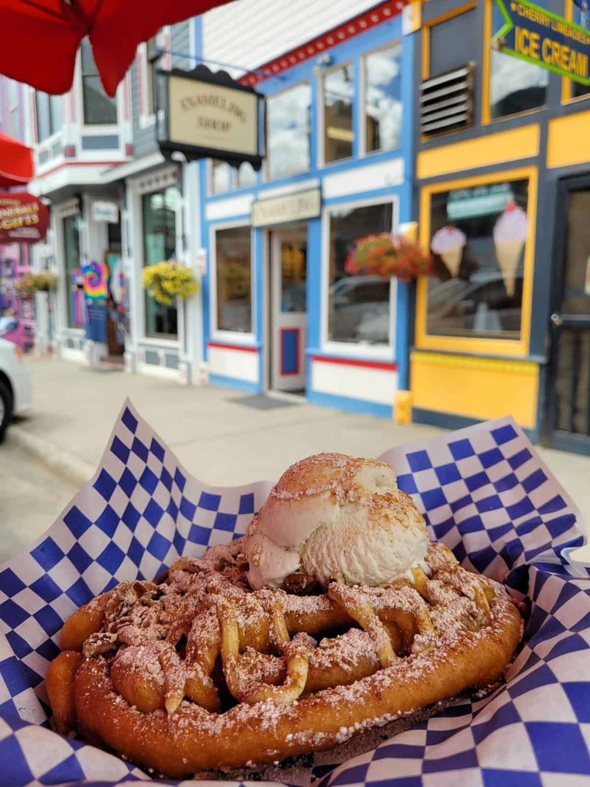 A close-up of a funnel cake topped with ice cream and powdered sugar on a checkered paper liner, with colorful storefronts in Silverton visible in the background