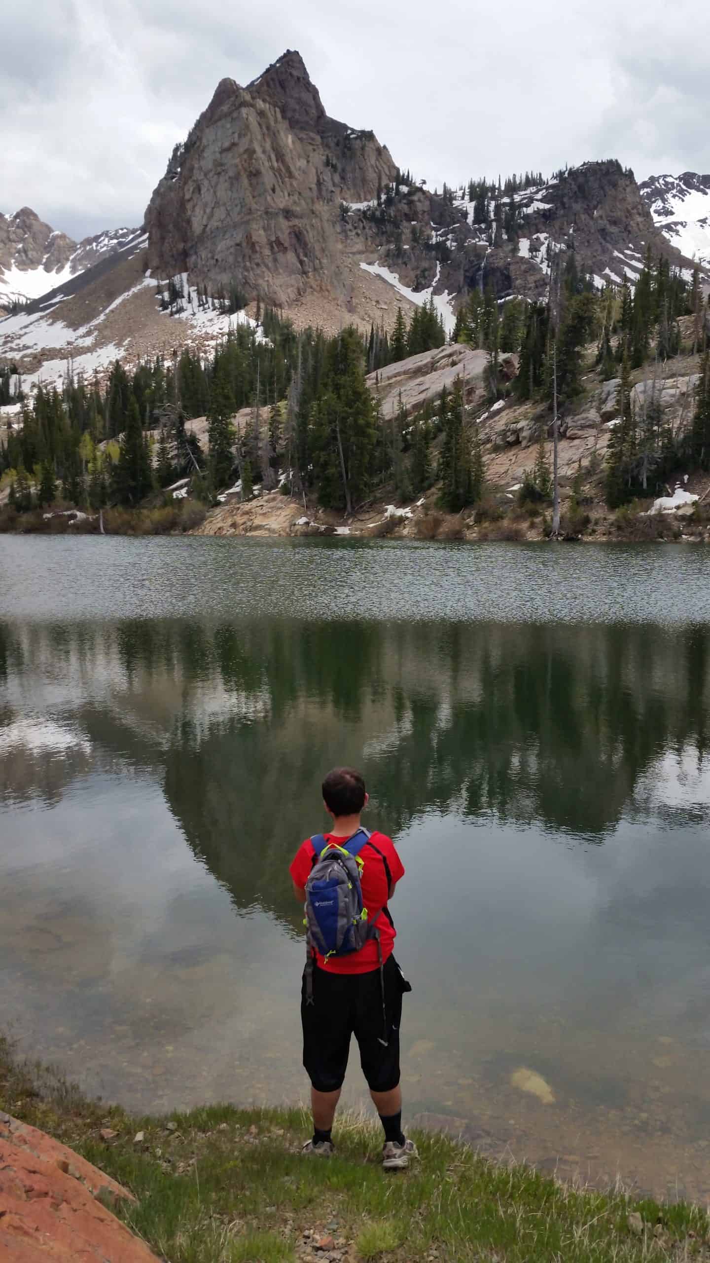A hiker viewing Sundial Peak reflecting in calm water on Lake Blanche in the early summer, under moody skies