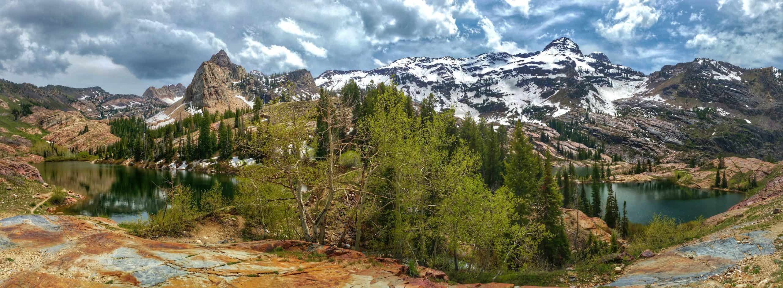 A panoramic view of Lake Blanche on the left and Lakes Florence and Lillian on the right, surrounded by rugged partially snowcapped mountains, forests, and colorful alpine terrain under a moody sky.

