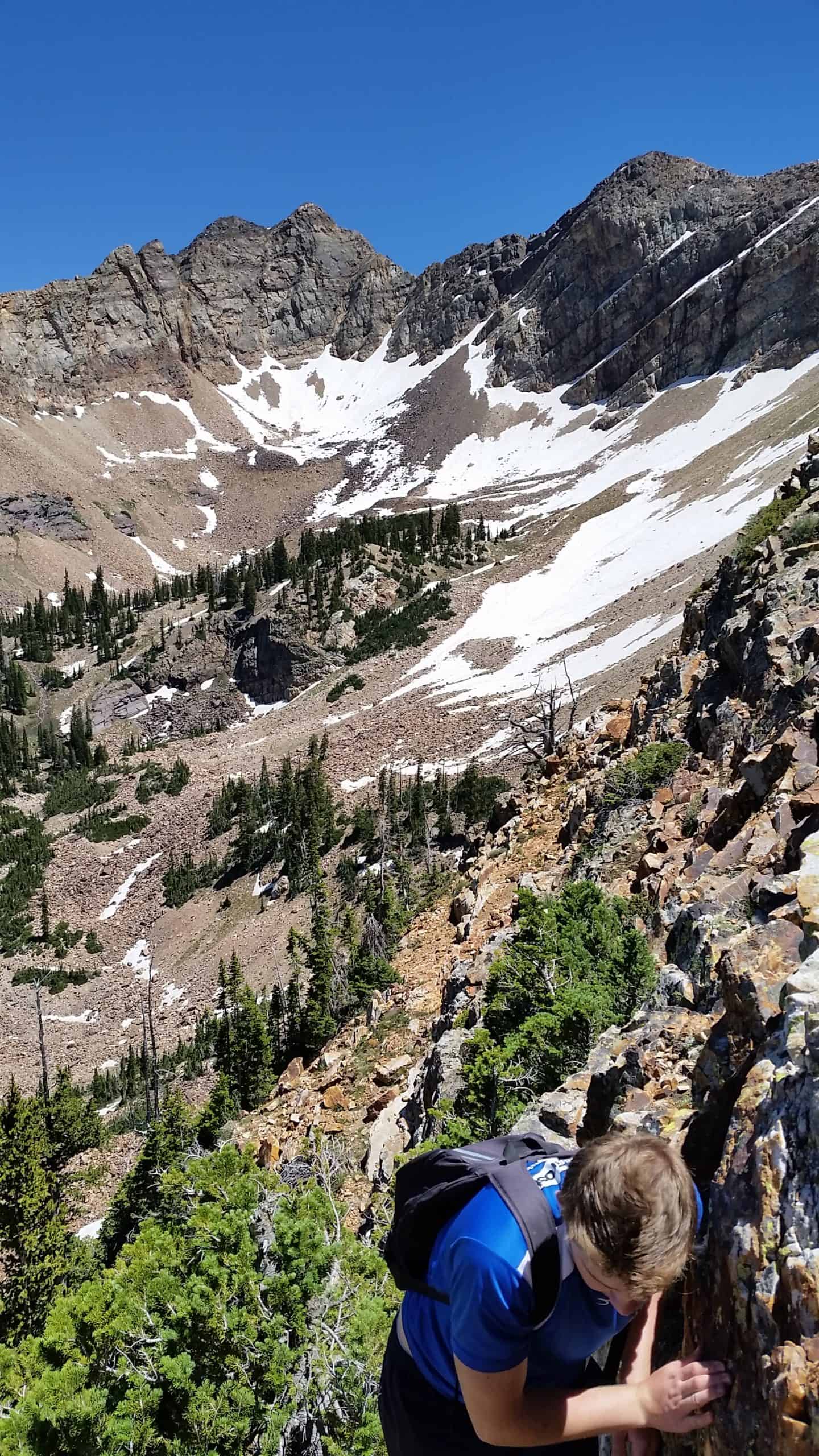 A hiker carefully climbs over rocky boulders, gazing down at a snow-dappled valley beneath a clear blue sky enroute to Sundial Peak

