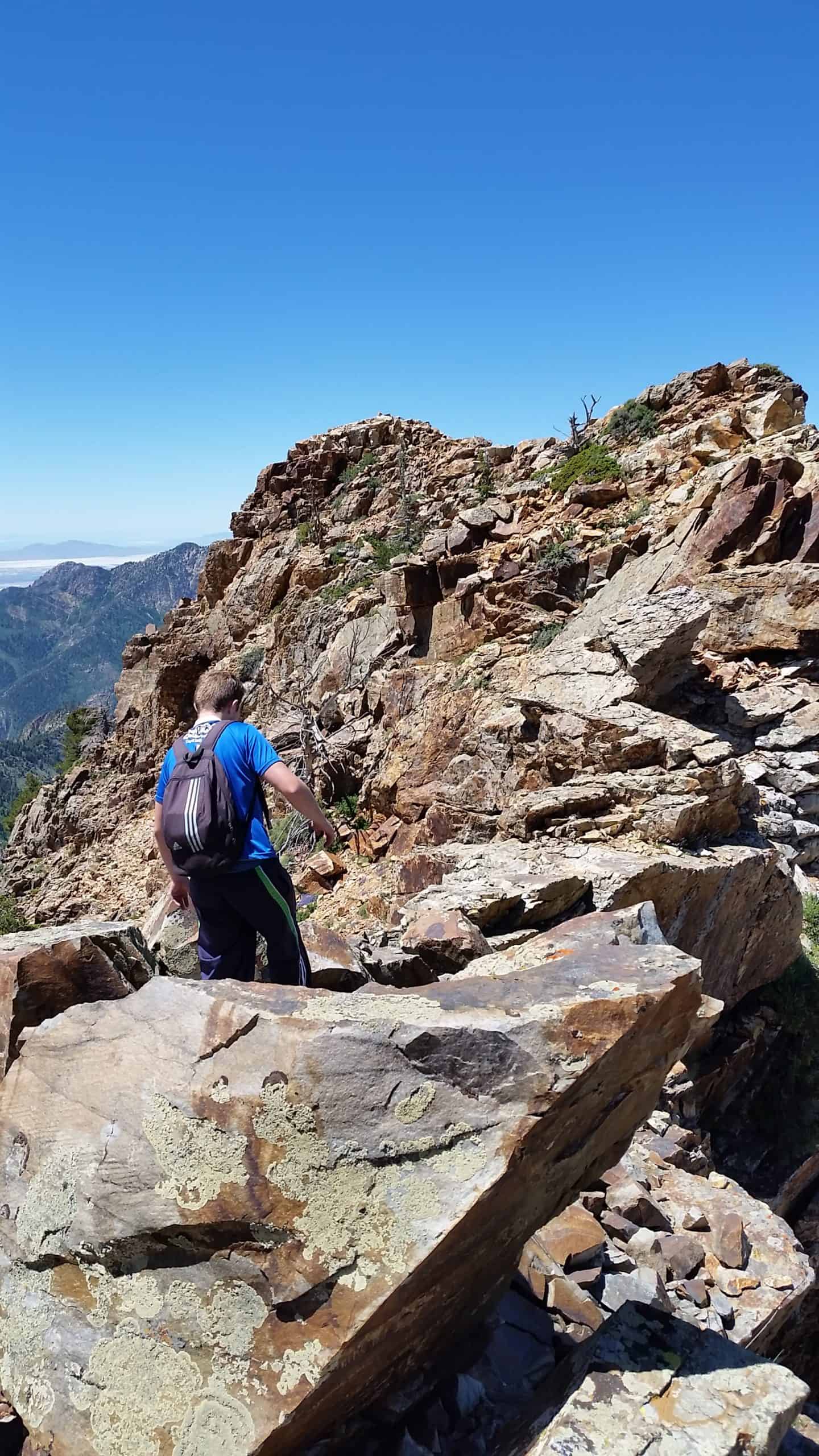 A hiker in a blue shirt walks carefully along a rugged rocky path towards the summit of Sundial Peak, on a sunny day, ascending through exposed mountain terrain.

