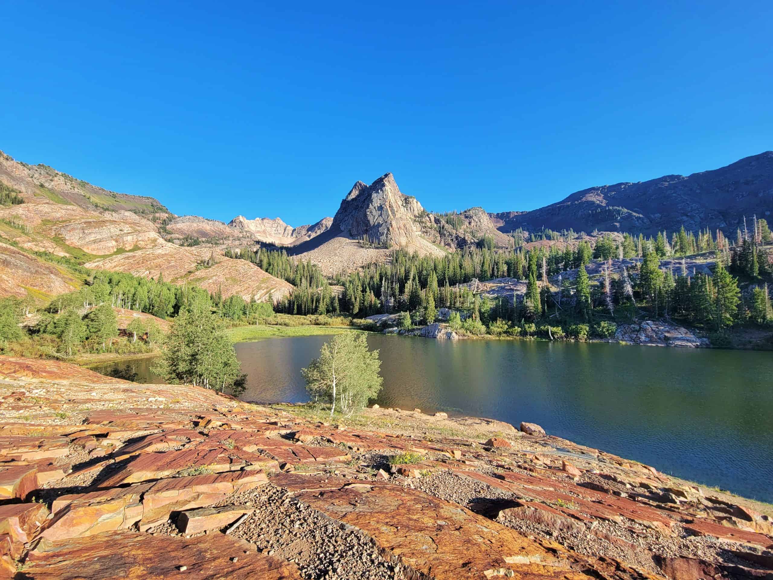  A slightly elevated view of Lake Blanche, with Sundial Peak framed in the center, surrounded by rocky slopes and trees under a clear blue sky