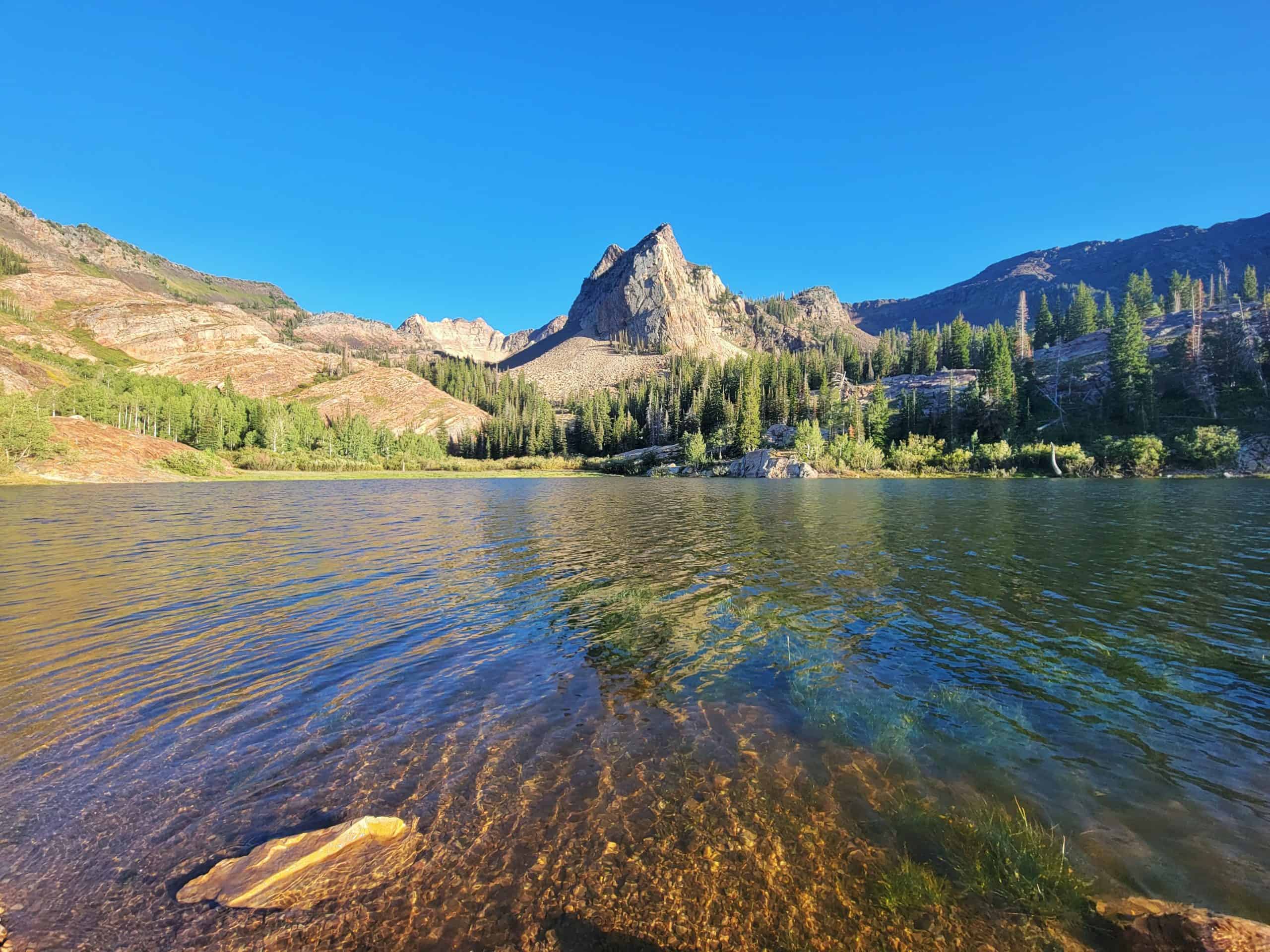 A serene view of Lake Blanche on a clear day, with calm waters reflecting the surrounding forested slopes and Sundial Peak in the distance.