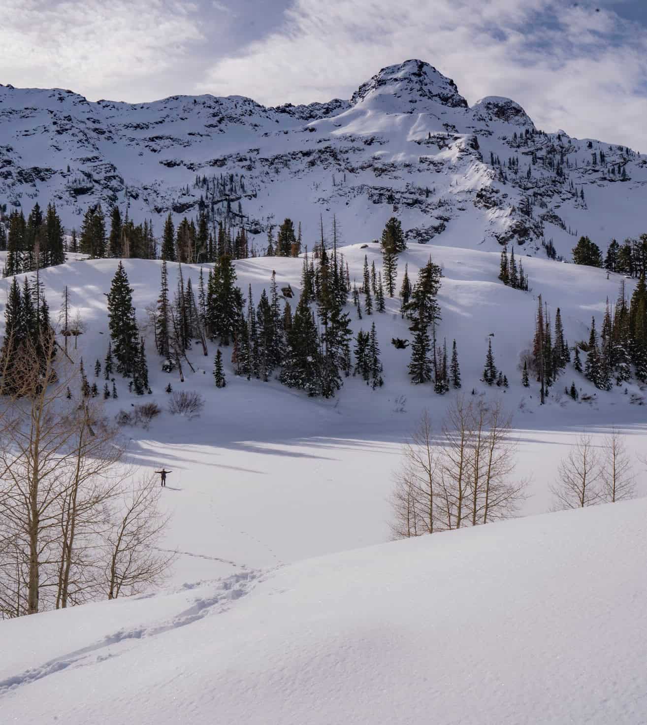 A hiker standing on a frozen Lake Blanche with tracks leading out to him and a snowy mountains landscape with pine trees
