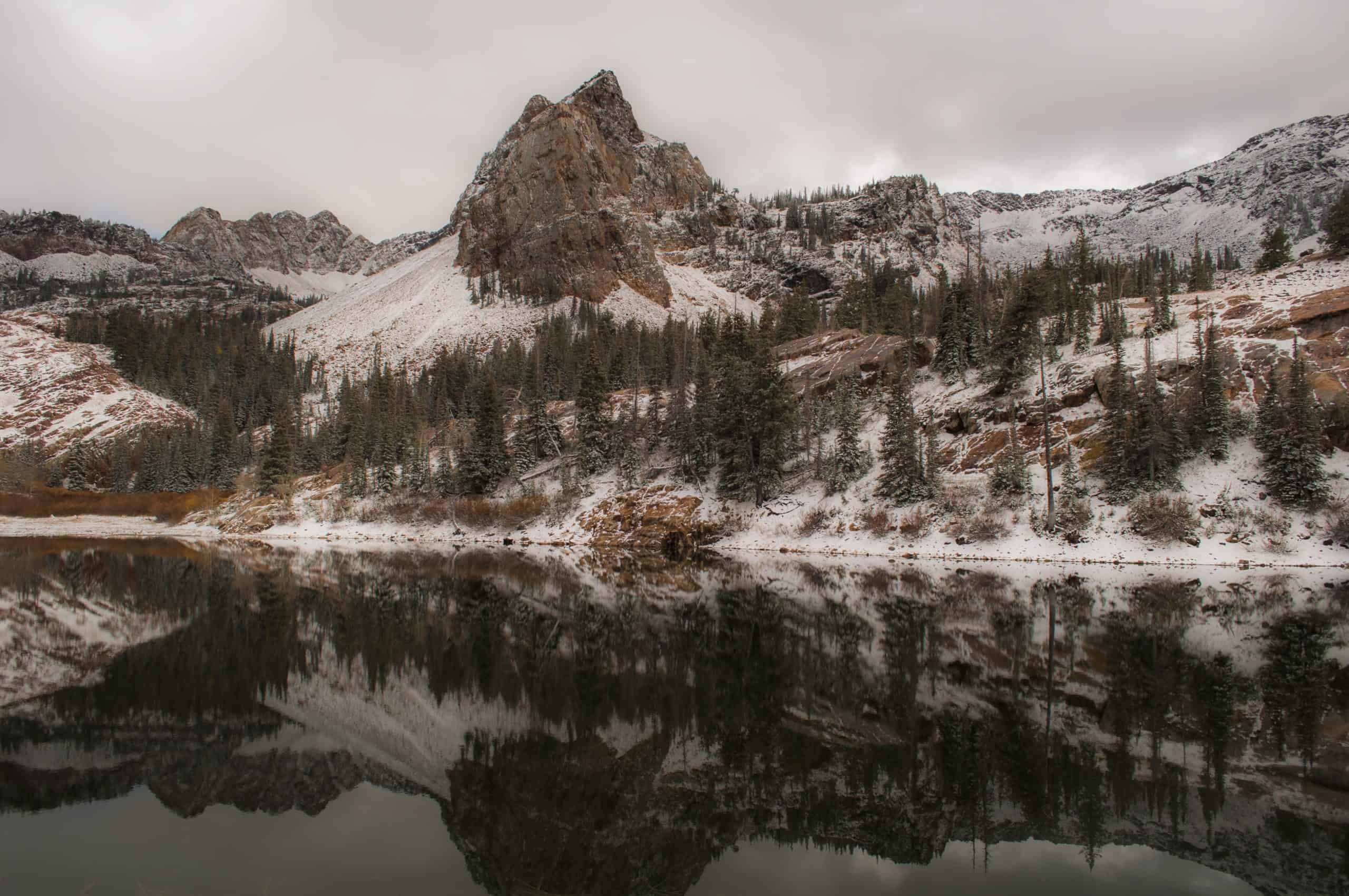 A gloomy view of Lake Blanche in fall, framed by hushed pine trees and soft mountain shadows beneath an grey-colored sky.

