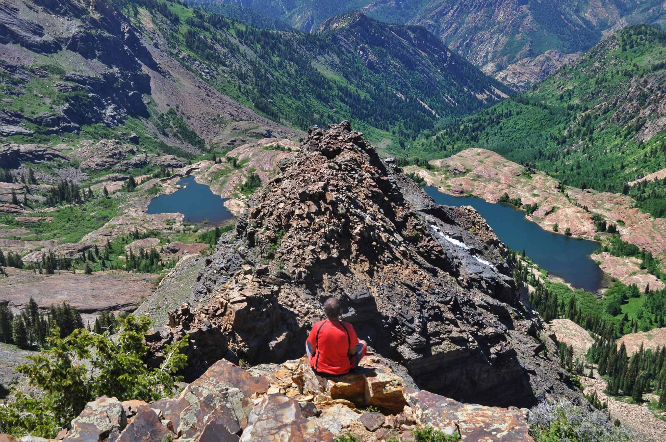 A serene view from Sundial Peak in the summer, with a hiker sitting on a rocky ledge overlooking two alpine lakes, including Lake Blanche, nestled among forested slopes and rugged mountains.

