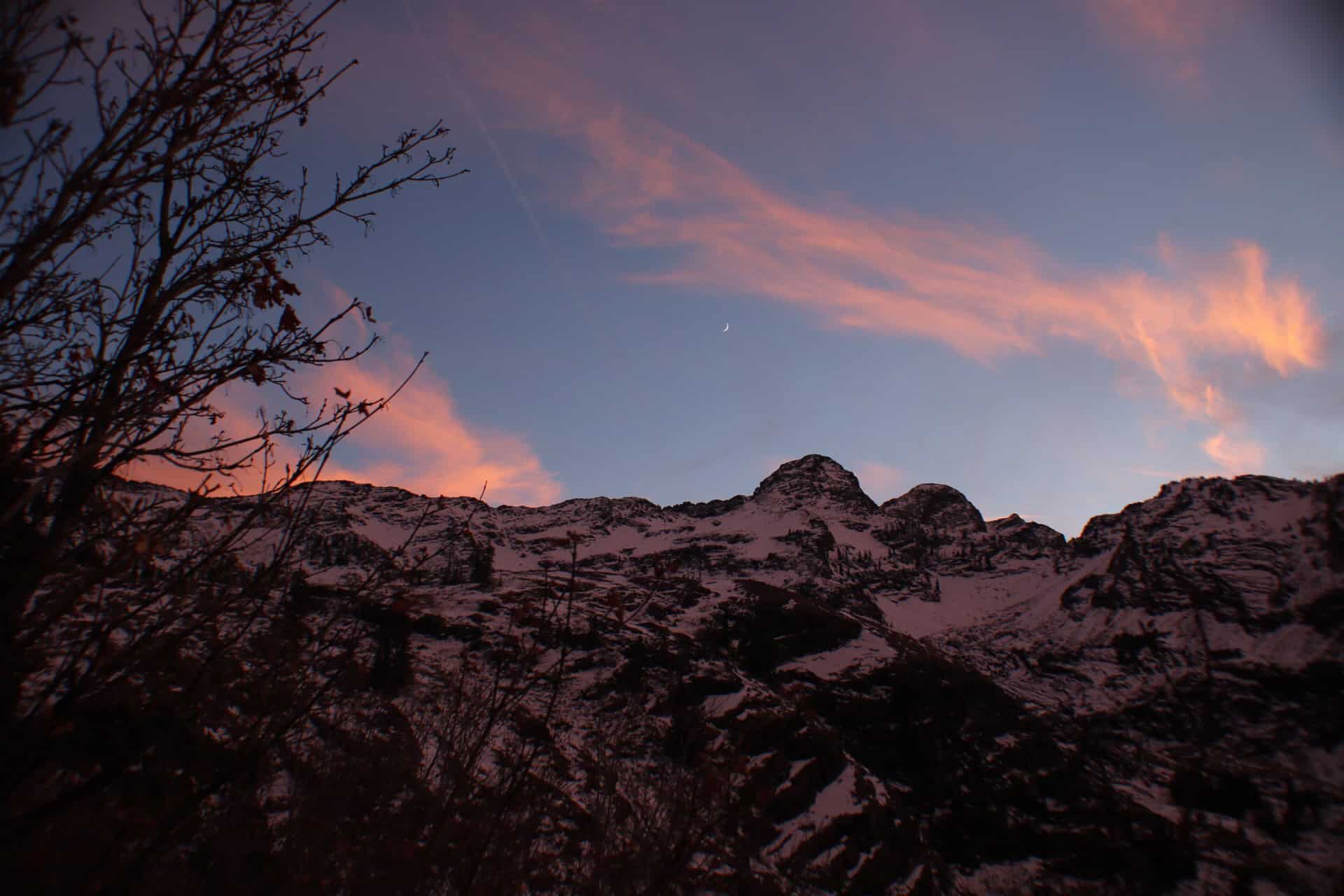 A scenic view of Dromedary and Sunrise Peaks from the Lake Blanche Trail, with snow-dusted ridgelines and tree branches silhouetted against a twilight sky at dusk.
